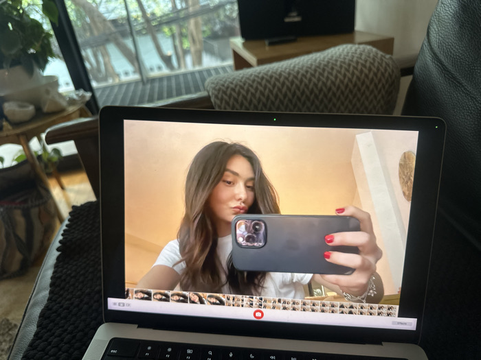 A silver-colored laptop is positioned on a dark, textured surface, likely a sofa or chair arm. The laptop screen displays a digital image of a young woman taking a selfie. The woman has long, dark, wavy hair and wears a white t-shirt, looking towards the camera with a slight pout. She holds a dark-colored smartphone, likely an iPhone, in her right hand, with her fingernails painted bright red. The background of the selfie shows a warm-toned interior wall, a white rectangular architectural element, and a circular decorative object. The laptop's display interface includes a filmstrip of smaller image thumbnails along the bottom edge and green indicator lights at the top of the screen bezel.
In the real-world background, to the left of the laptop, a light-colored wooden side table is visible, holding several white bowls or decorative items and a green potted plant. Beyond this, a large window or glass door offers a view of an outdoor area with lush green trees and a dark metal balcony railing. To the right of the laptop, a patterned cushion and a dark leather sofa are partially visible. A dark rectangular object, possibly a speaker, sits on a wooden surface behind the cushion. The scene is illuminated by natural light entering through the window. This setting is a residential interior in Mexico City, Mexico.