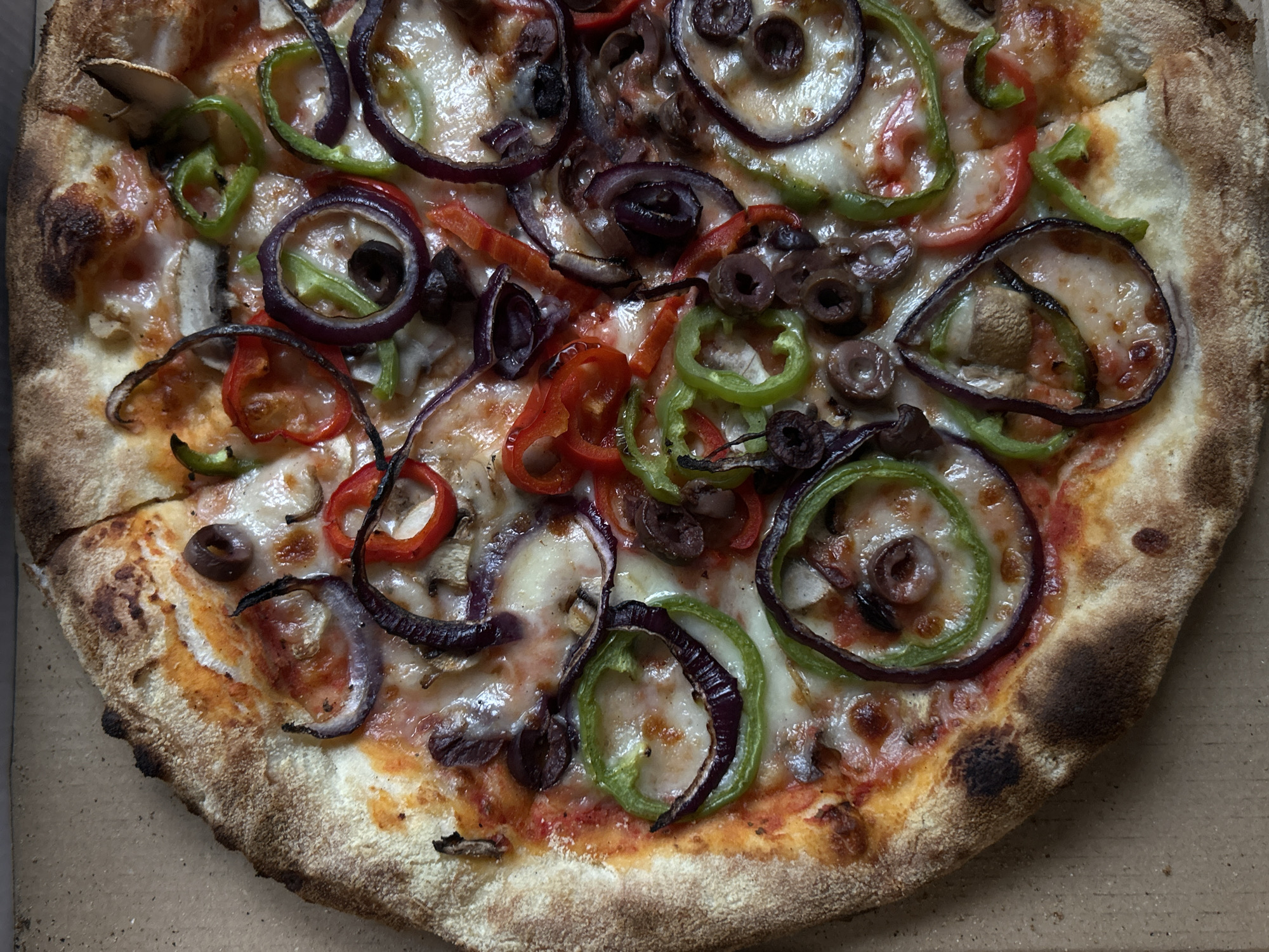 An overhead, close-up view captures a freshly baked pizza resting within a simple, light brown cardboard box. The pizza is generously adorned with a colorful array of toppings over a base of melted, bubbly cheese and red tomato sauce.
Prominent among the toppings are thinly sliced red onions, some maintaining their distinctive purple rings, while others appear slightly separated and caramelized. Bright green bell pepper slices, mostly in rings, are scattered across the surface, providing a vibrant contrast. Sliced black olives are visible throughout the pizza, alongside occasional slices of mushrooms. The cheese is thoroughly melted, creating a glossy, appetizing texture with some slightly browned areas, indicating even baking.
The pizza features a substantial, puffed-up crust that is golden brown with varying degrees of charring, suggesting high-temperature baking, possibly in a wood-fired oven. The crust appears artisanal, with visible air bubbles and a rustic texture. One slice of the pizza appears to have been slightly moved or cut, creating a small gap near the left side.
The image focuses entirely on the pizza, showcasing its fresh and appetizing appearance. No specific time of day or weather conditions can be discerned. There is no visible text or branding on the pizza or the cardboard box.