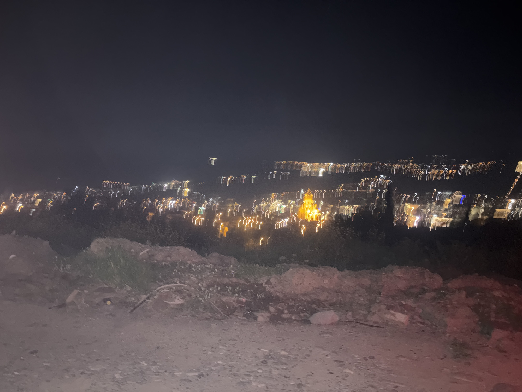 Here is a concise description of the scene:
The image presents a blurred night view of the city of Tbilisi, Georgia. The foreground consists of a rocky, dirt-covered slope with sparse vegetation. Beyond the slope, the lights of the city are visible, appearing as elongated streaks due to motion blur. There are no visible people or interactions. The environment is dark and appears to be an elevated vantage point overlooking the city.