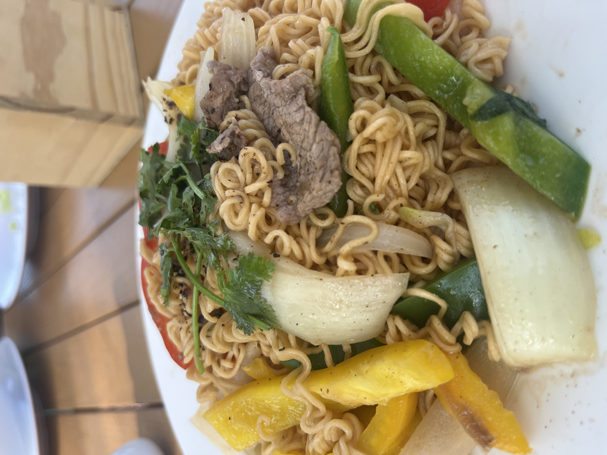 A top-down shot depicts a plate of stir-fried noodles with vegetables and meat. The noodles are curly and light brown. The vegetables include yellow and red bell peppers, green beans, onions, and leafy greens, possibly cilantro. Slices of meat, likely beef, are also visible. The plate is white and placed on a wooden surface. The location is unknown.