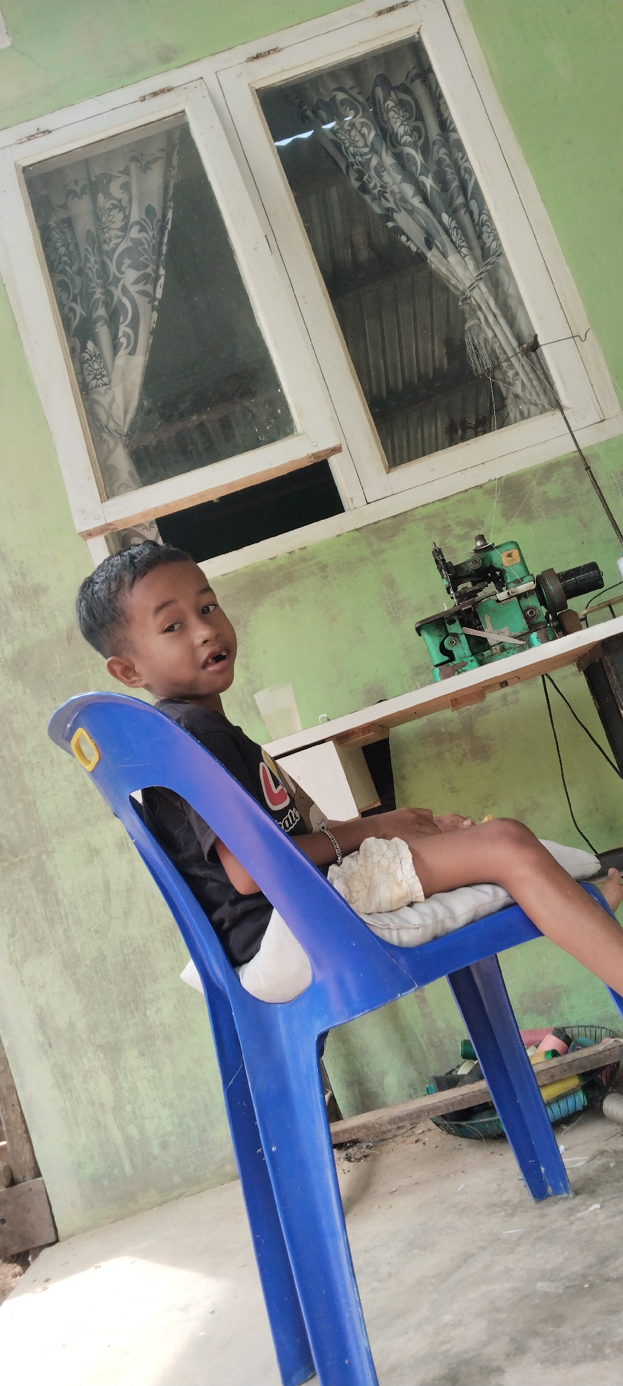 A young boy of Southeast Asian descent, with dark hair and an open mouth, sits on a blue plastic chair in an outdoor setting, likely Meunasahmee, Indonesia. He is wearing a black t-shirt with a red logo and light-colored shorts. A thin silver bracelet is visible on his left wrist. The chair has a yellow accent on its handle.
Behind the boy, a white table is visible, with a green sewing machine positioned on it. A tall, thin pole extends upwards from the table, with threads hanging from it. In the background, a light green wall serves as a backdrop. Above the wall, a large window with white framing is present, covered by patterned curtains in shades of black and white. A part of the window opening appears to be covered by a wooden plank.
The floor is made of concrete and appears to be partially covered with debris. To the right of the chair, beneath the table, a basket filled with spools of thread in various colors, including pink, yellow, and blue, is visible. The overall environment suggests a domestic or workshop setting.
