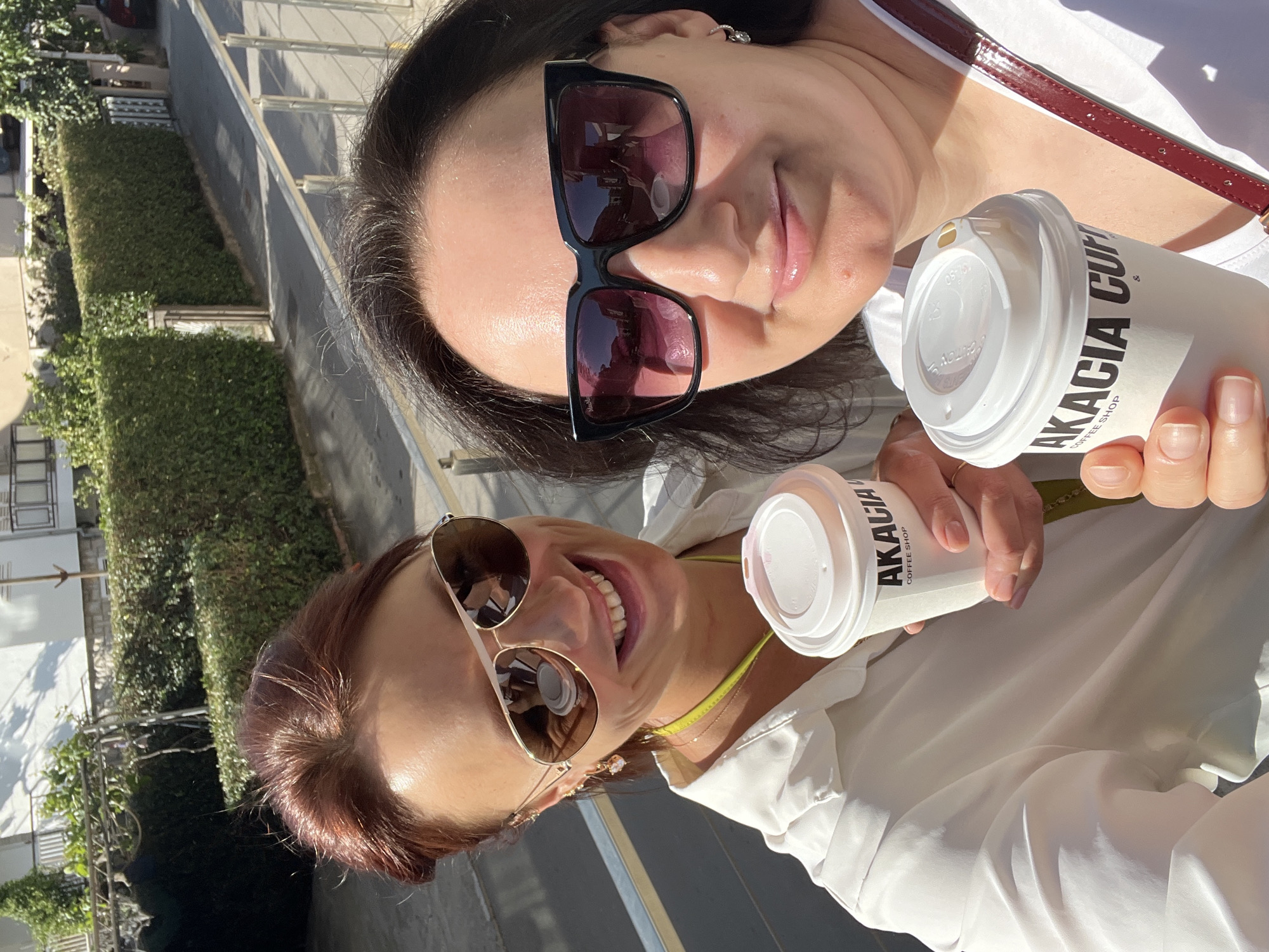 Two women are posing for a selfie outdoors. The woman in the foreground has auburn hair pulled back, sunglasses, a white blouse, and a neon green strap from an unknown object. She is holding a white paper coffee cup labeled "AKACIA COFFEE SHOP." The woman in the background has dark hair, sunglasses, a white top, and a red purse strap. She is also holding a "AKACIA COFFEE SHOP" cup. The background includes a building, greenery, and a sidewalk. The location appears to be in an urban setting. Akacia is a coffee shop located in Budva, Montenegro, indicating the photo was taken in Budva.