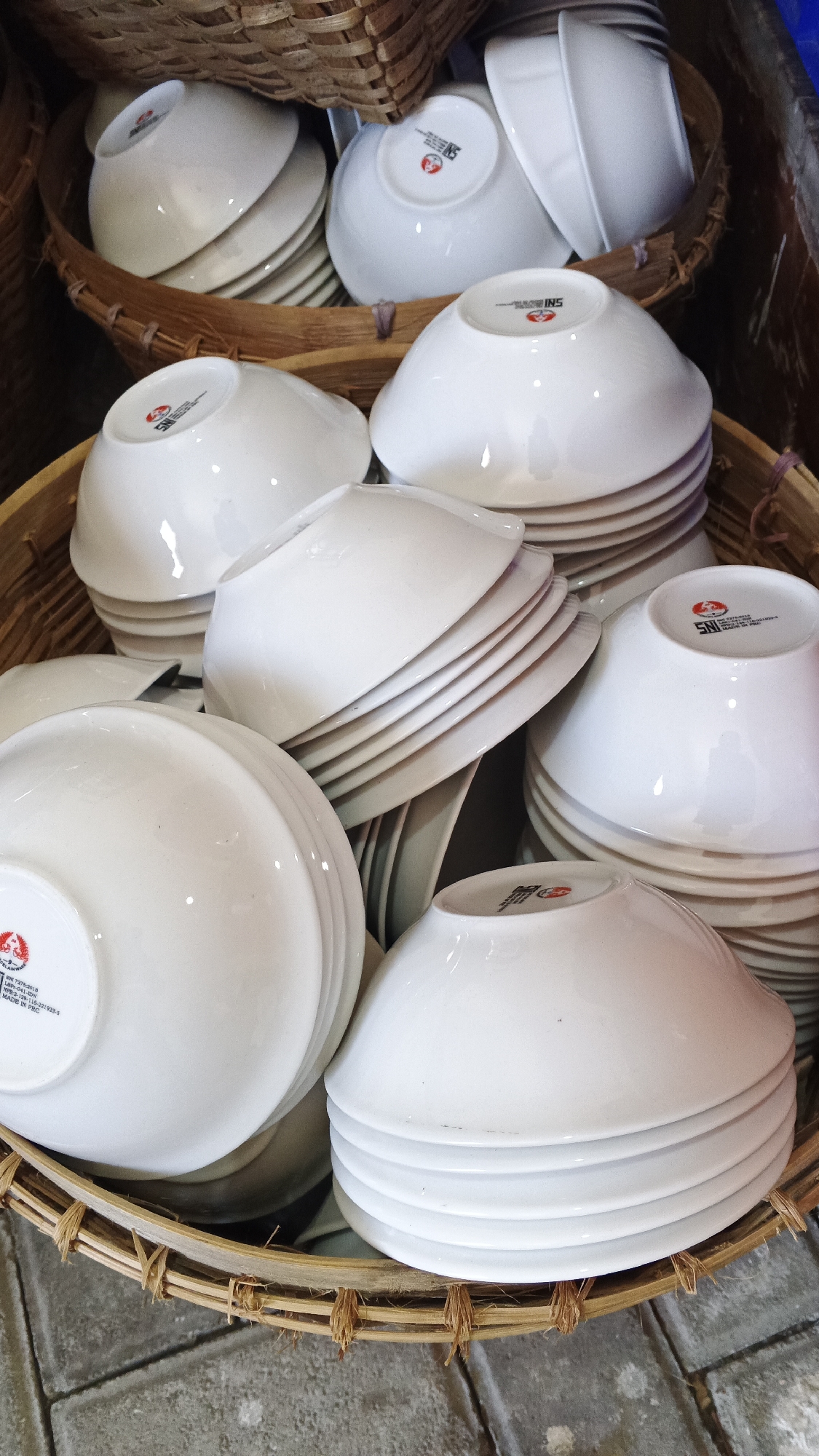 A high-angle, close-up shot shows a woven basket filled with stacked white ceramic bowls. The bowls are smooth and have a slightly glossy finish. Some of the bowls have a red and white logo with the text "SNI" printed on the inside bottom. The basket is made of thin, light brown reeds that are woven together and secured with visible twine. The basket sits on a grey, textured surface that appears to be cobblestone or rough concrete. The lighting is natural and slightly diffused, casting subtle shadows. The image is sharp and in focus. The scene is likely from a market or stall in Yogyakarta, Indonesia, where such items are commonly sold.
