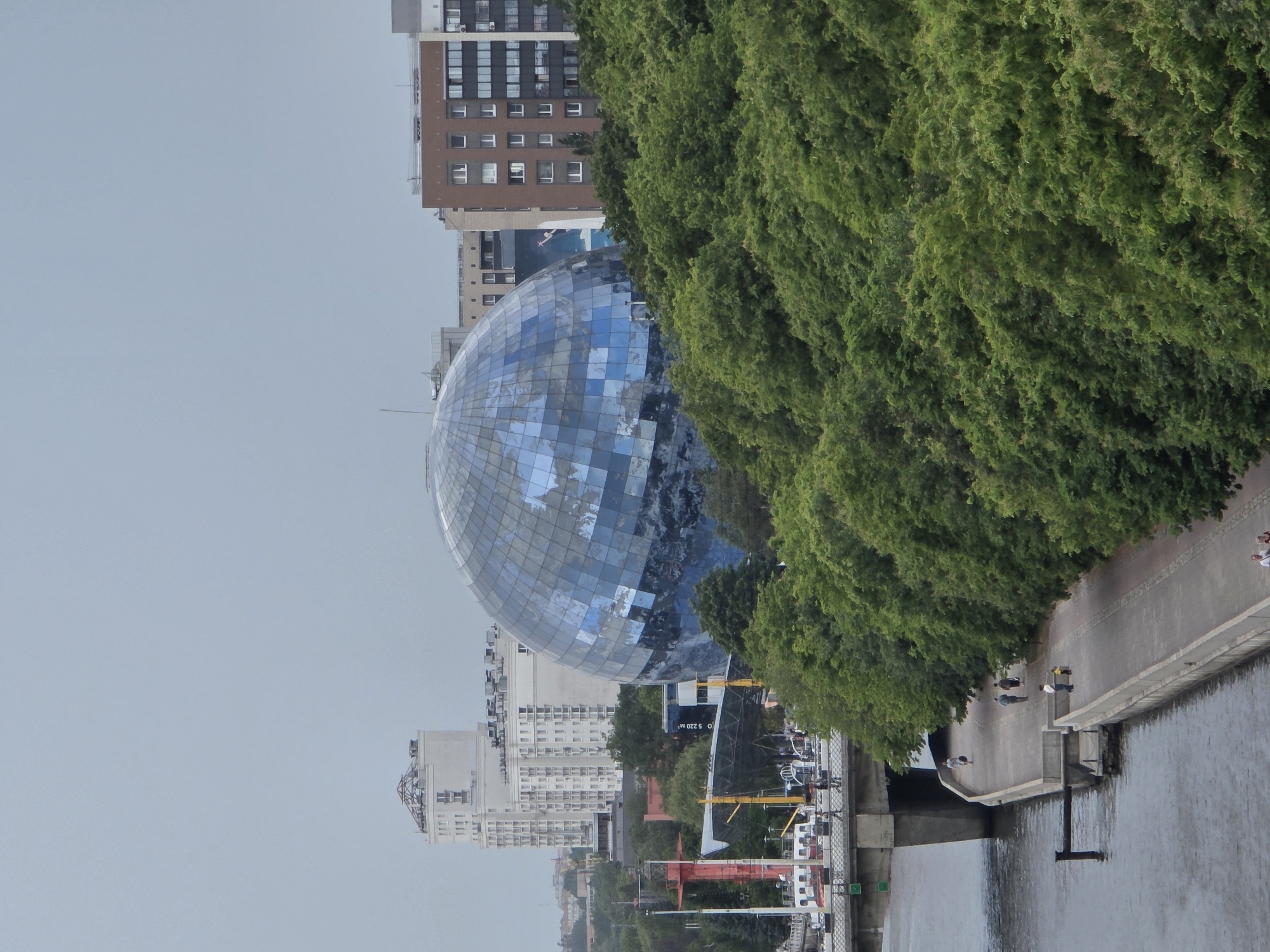 An elevated, oblique view depicts a section of Kaliningrad, Russia, under a uniformly overcast, light-grey sky. The central feature is a large, spherical building with a tessellated exterior of reflective blue and grey panels, known as the "Planet Ocean" exhibit of the Museum of the World Ocean. To the left and behind this sphere, several tall, light-colored residential buildings are visible, the tallest featuring a prominent, complex metal structure on its roof. To the right of the sphere, a multi-story building with a reddish-brown facade and numerous windows stands, with a large mural of a human figure visible on a building further back.
A dense expanse of green deciduous trees occupies the mid-ground and foreground, extending parallel to the river. The Pregolya River flows along the left side of the frame, presenting a calm, dark-grey surface. A concrete bridge crosses the river, with two individuals visible walking across it. Adjacent to the river, a paved promenade, constructed from light-colored concrete, extends into the foreground. On this promenade, several people are observed: one individual walking alone in the distance, a pair walking together, and closer to the foreground, an adult with a small child in a yellow top. A dark, horizontal pipe-like structure extends from the promenade into the river. On the riverbank near the bridge, a red, slender, lighthouse-like structure is visible, alongside portions of a large, dark, ship-like exhibit and a white moored boat, all associated with the Museum of the World Ocean complex. The scene captures a modern urban waterfront on an overcast day.