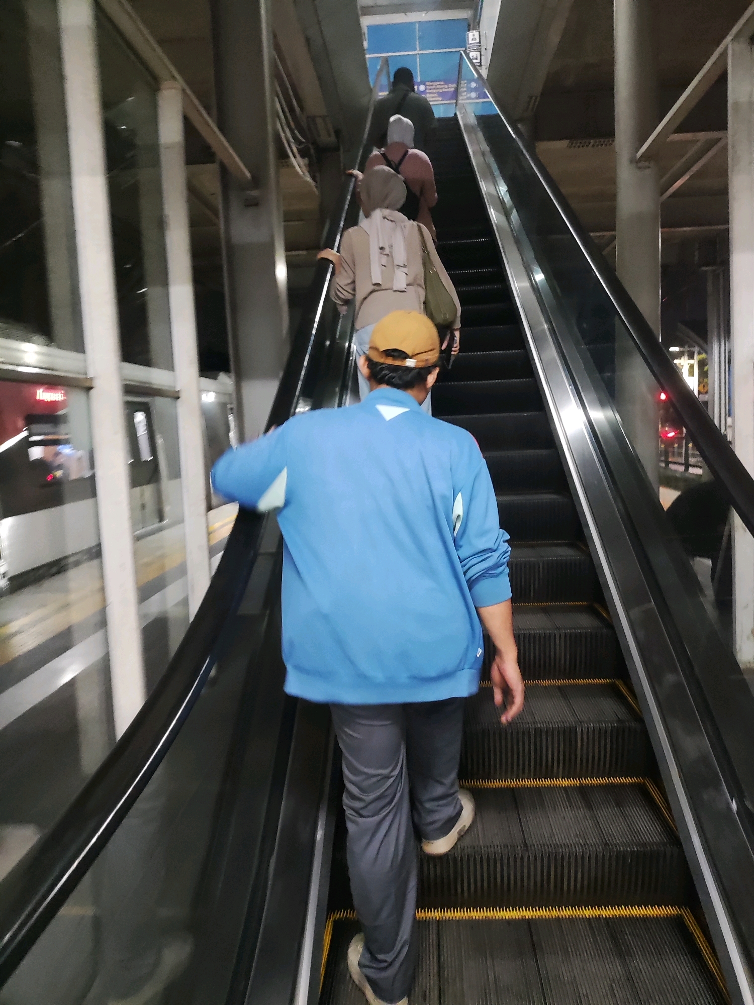 The image shows an upward-moving escalator at a transportation hub, likely a train station or airport, in East Jakarta, Indonesia. Several individuals are ascending the escalator, facing away from the camera. In the foreground, a person wearing a blue jacket and a tan cap is ascending. Behind them, other individuals are visible, some wearing head coverings. The environment appears to be an indoor or covered outdoor area with a modern aesthetic, featuring glass panels and concrete structures. The escalator steps are dark gray with yellow safety markings. The lighting suggests it might be evening or an area with artificial illumination.