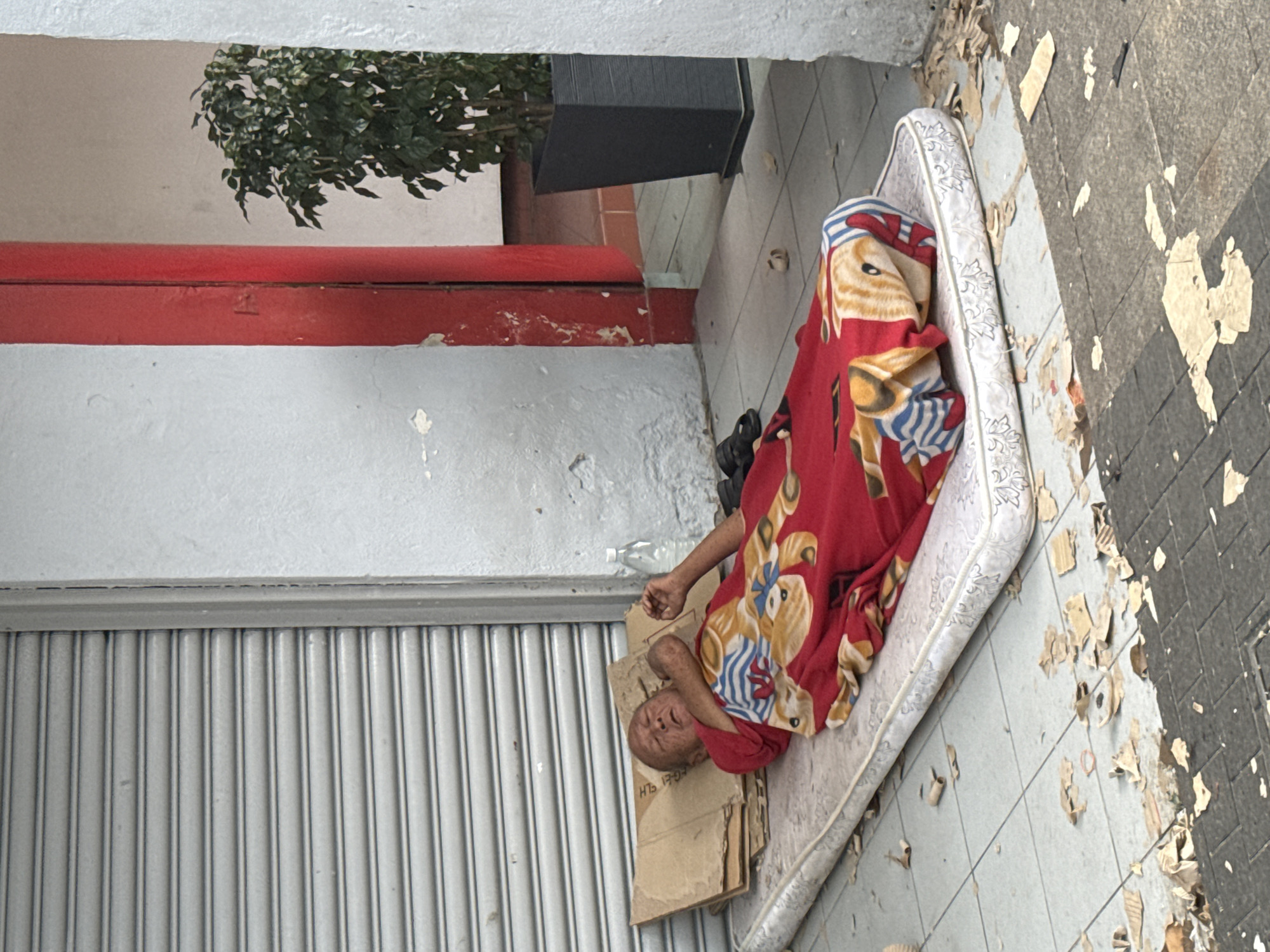 An unhoused individual sleeps on a mattress covered with a red blanket on a tiled sidewalk next to a building with a gray rolling metal door and a red-and-white trim. Cardboard pieces provide additional support. Debris is scattered on the sidewalk. The location is likely in Kuala Lumpur, Malaysia, as specified.
