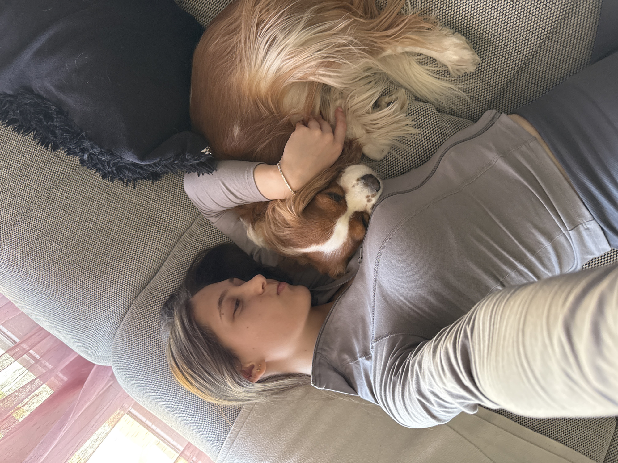 A woman and a dog are resting on a grey, textured sofa indoors, viewed from an overhead perspective. The woman is lying on her back, facing left, with her eyes closed or partially closed, suggesting she is asleep or deeply relaxed. She has light brown hair with blonde highlights and is wearing a plain grey, long-sleeved top. A small earring is visible in her right ear, and a simple silver bracelet adorns her left wrist.
A brown and white dog, possibly a Cavalier King Charles Spaniel, is curled up against the woman's chest and shoulder, also appearing to be asleep. The dog has long, fluffy fur. The woman's left arm is gently draped over the dog's back, indicating a close bond.
Behind the woman's head is a dark, possibly black, cushion or blanket with a fringed edge. To the lower left of the frame, a window allows natural daylight to enter, partially covered by pink, sheer curtains or blinds. The lighting suggests it is daytime. The scene conveys a peaceful and affectionate moment of rest or napping between the woman and her pet within a comfortable home setting. No visible text is present in the image.