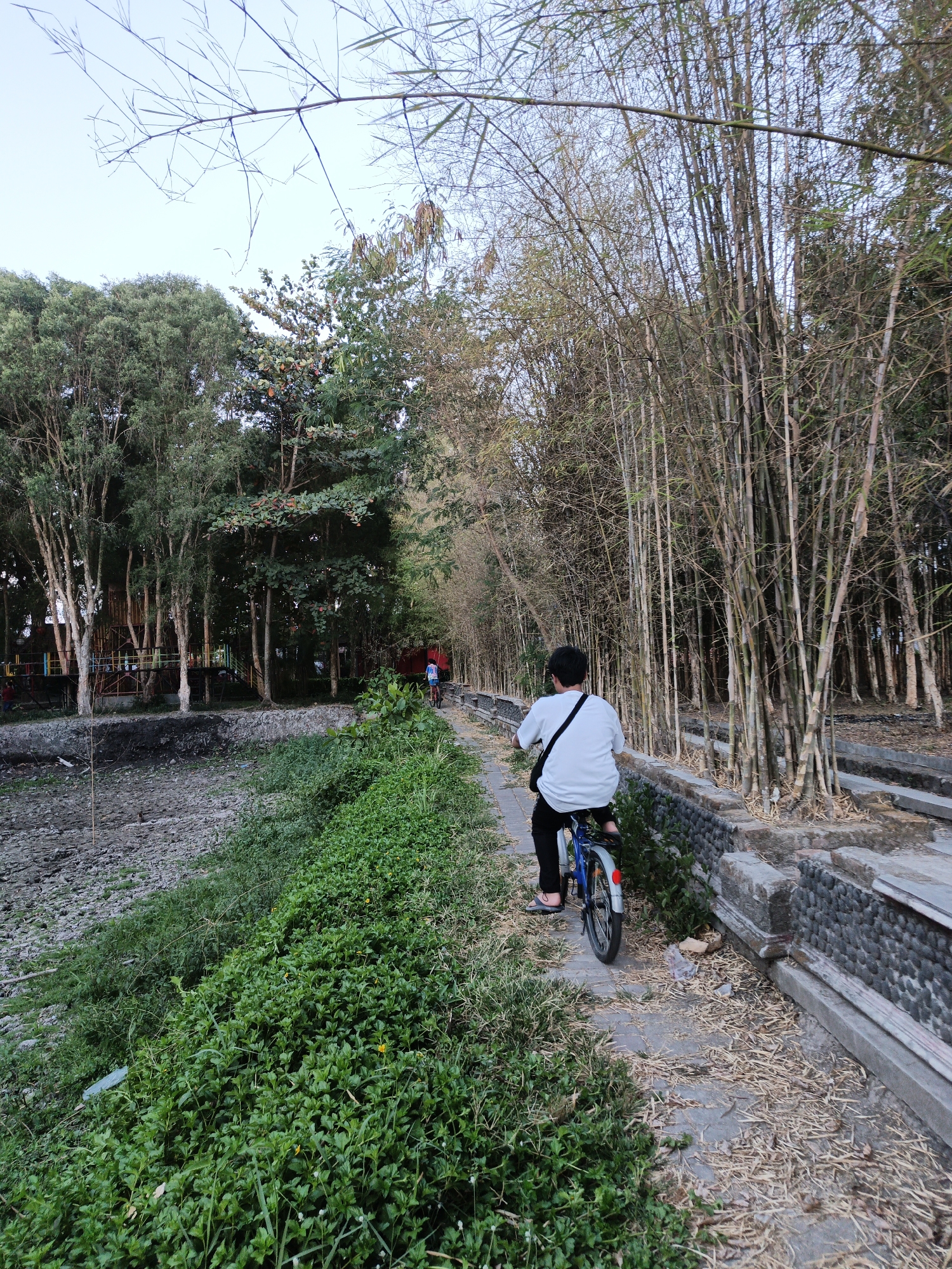 In Klaten, Indonesia, a person on a bicycle rides down a paved path bordered by lush green vegetation on the left and a dense bamboo forest on the right. The path is also lined with a decorative stone wall. In the background, a playground structure is visible among trees. The sky is overcast.