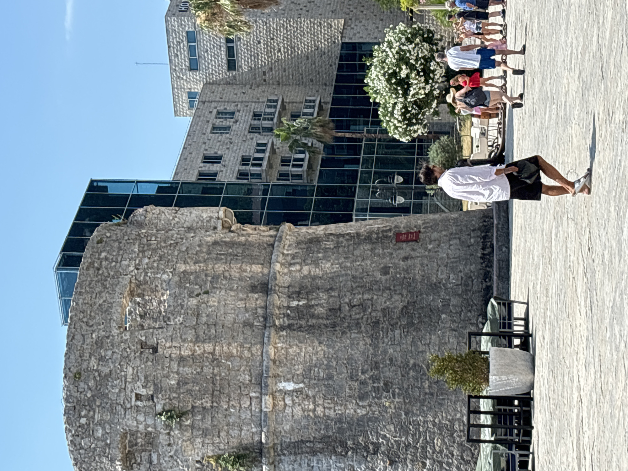 The image depicts a sunny day in the city of Budva, Montenegro. In the foreground, a man in a white shirt and black shorts walks across a paved plaza. Behind him, several people are visible, some walking and others standing. To the left, a large, ancient stone tower stands prominently. In the background, a modern hotel building with a glass facade and stone exterior rises above the old tower. A flowering tree adds a touch of color to the scene, and palm trees are also present. The overall impression is of a historical setting blended with contemporary architecture.