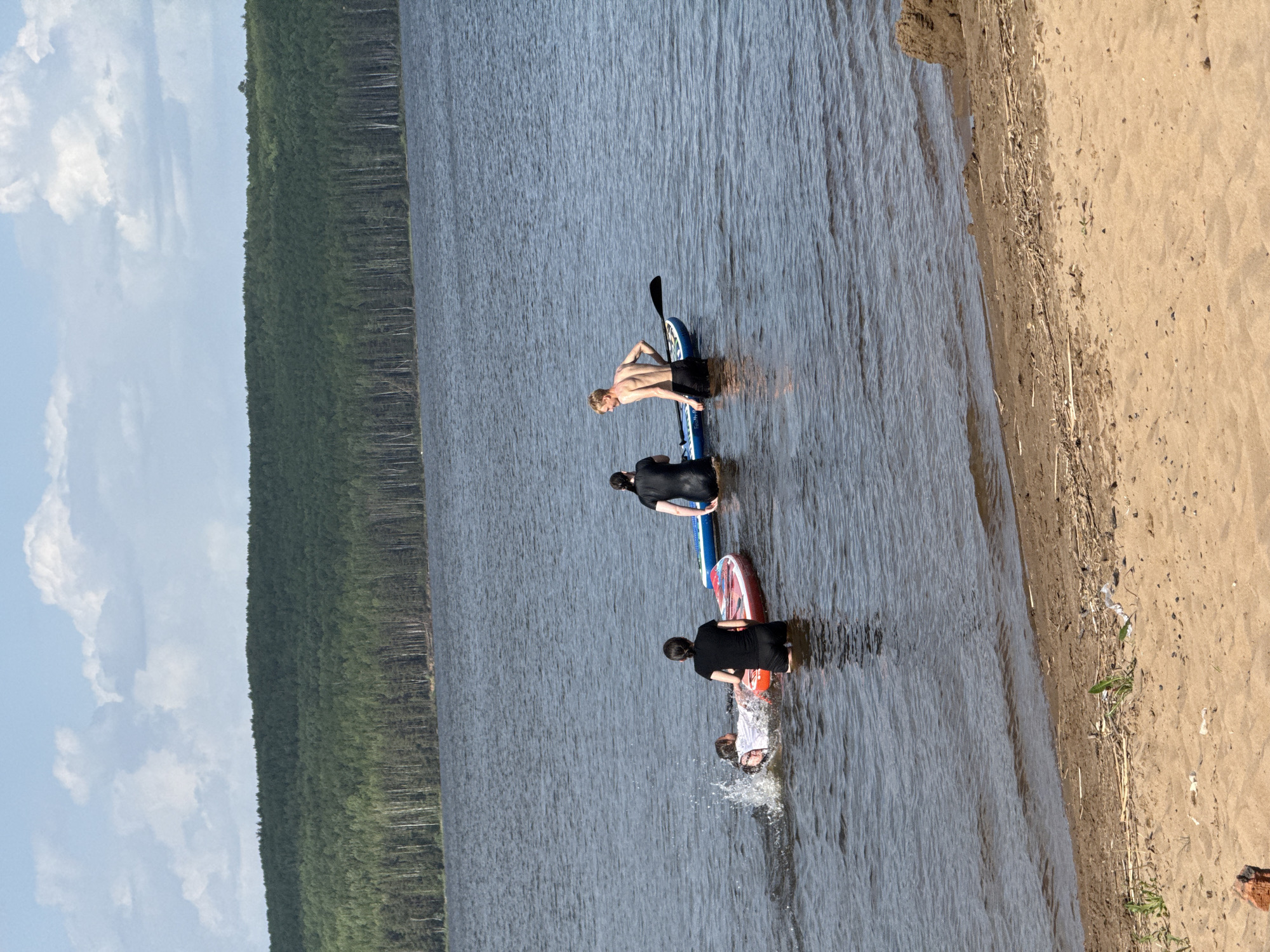 The image shows a group of people on paddleboards in a body of water near a beach. The location appears to be in a natural environment, with a forest visible on one side of the water. Three paddleboards are in the water, with people standing on them. The person farthest from the camera is shirtless, while the other two are wearing dark clothing. A fourth person is in the water with a red paddleboard and looks like they are falling in the water. The beach is sandy, with some driftwood and debris visible along the waterline. The sky is partly cloudy.