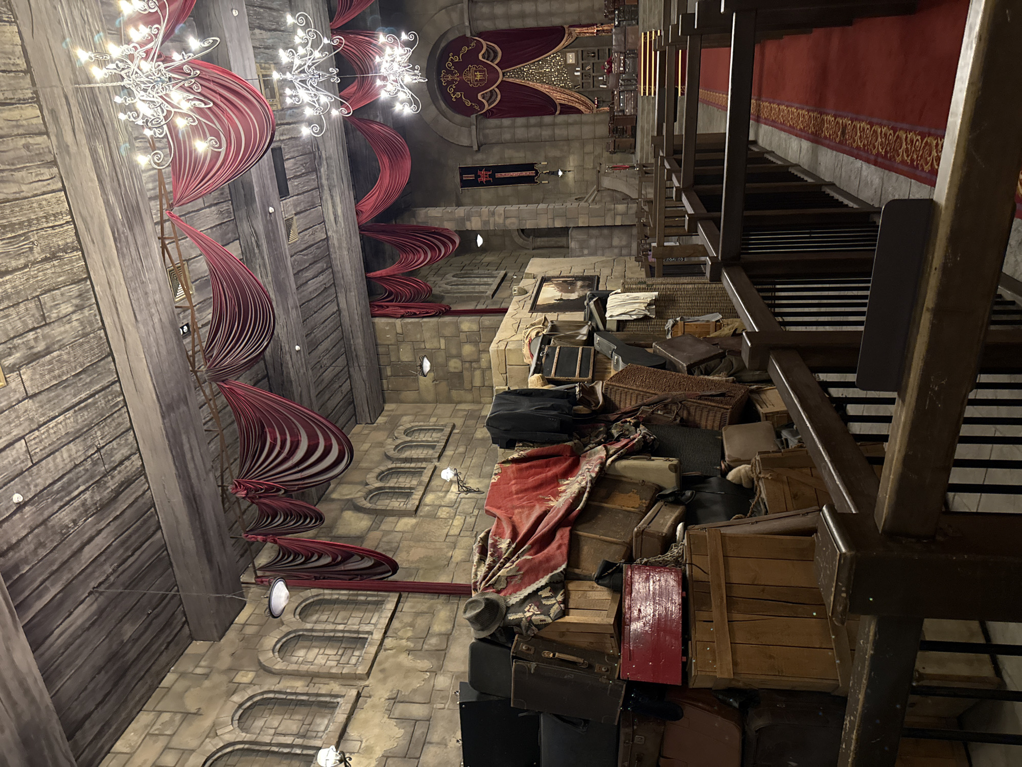 The image displays an elaborate, dimly lit indoor hall, resembling a grand castle interior or a fantasy-themed attraction. The perspective is tilted, showing the scene rotated clockwise.
The high ceiling on the left features dark wooden planks and multiple ornate, white chandeliers with numerous small lights, casting a sparkling glow. Rich red draped fabric hangs decoratively from the ceiling and walls in swirling patterns, complementing the light-colored stone block walls below. These walls include arched indentations and small, embedded light fixtures.
On the right side, a substantial, cluttered pile of antique-style luggage dominates the foreground, consisting of various suitcases, trunks, and wooden crates. A grey fedora hat rests on the pile, which is partially covered by a red and gold patterned fabric. Adjacent to the luggage is a dark wooden structure, appearing to be a steep staircase or ramp, lined with a red carpet runner that has a gold decorative border.
In the background, a large red and gold tapestry with a detailed emblem hangs on the far wall. Below it, a dark banner with a crest and some visible, but mostly illegible, text (possibly "HOGWARTS") is displayed. The overall lighting is artificial and dramatic, creating a mysterious and immersive atmosphere. No people are visible, and there is no direct activity depicted, but the stacked luggage strongly suggests a theme of travel, arrival, or departure within this grand setting. The architecture and decor cues point to a location designed to evoke a sense of history, fantasy, or magic.