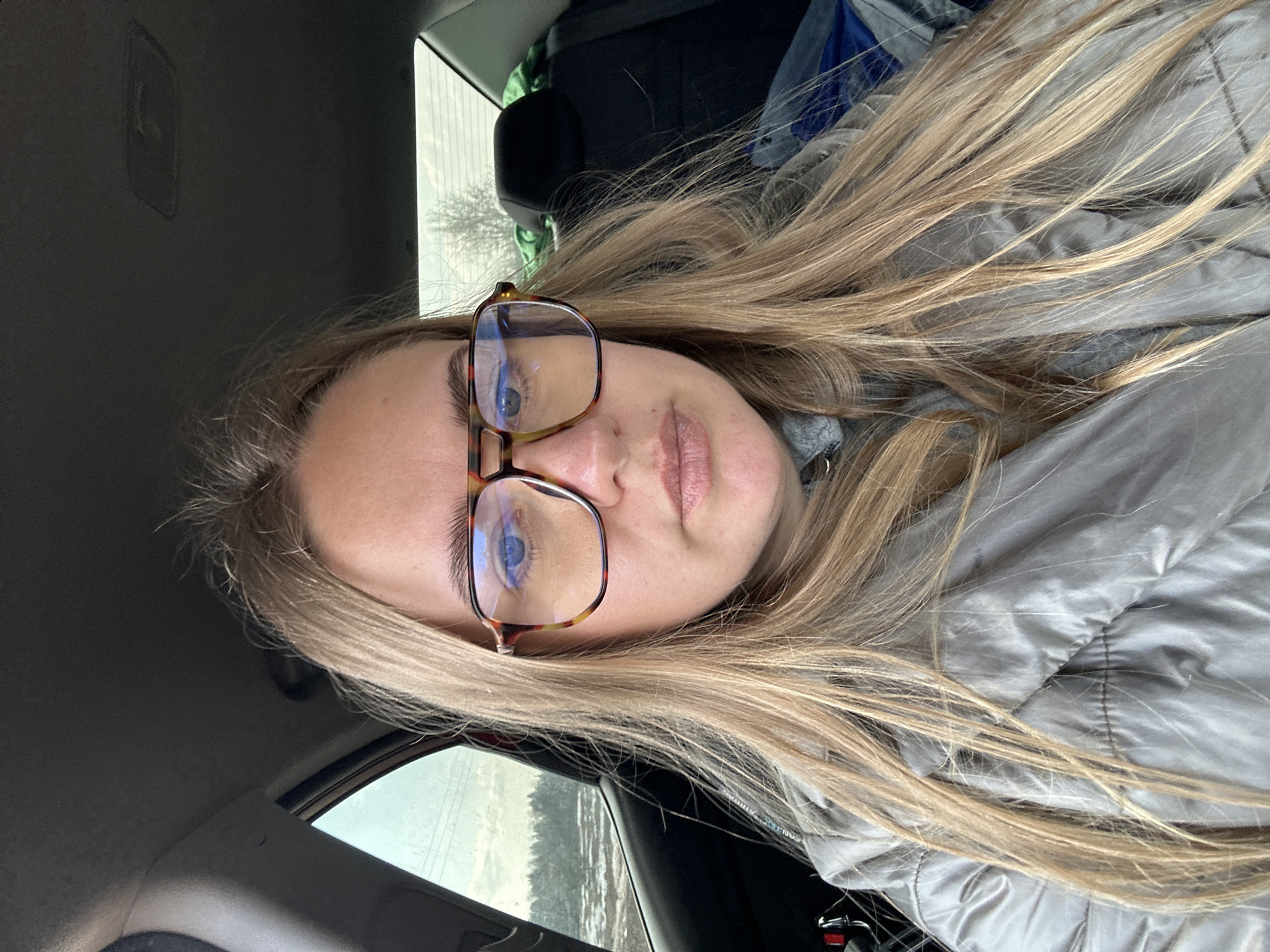 A young woman with long blonde hair and wearing tortoiseshell-framed glasses is in the passenger seat of a car. She is wearing a light grey puffy jacket. Outside the car window, the landscape appears to be a snow-covered field with sparse trees. The image appears to be taken from inside the vehicle. The location is likely Ural'skoe, Russia.