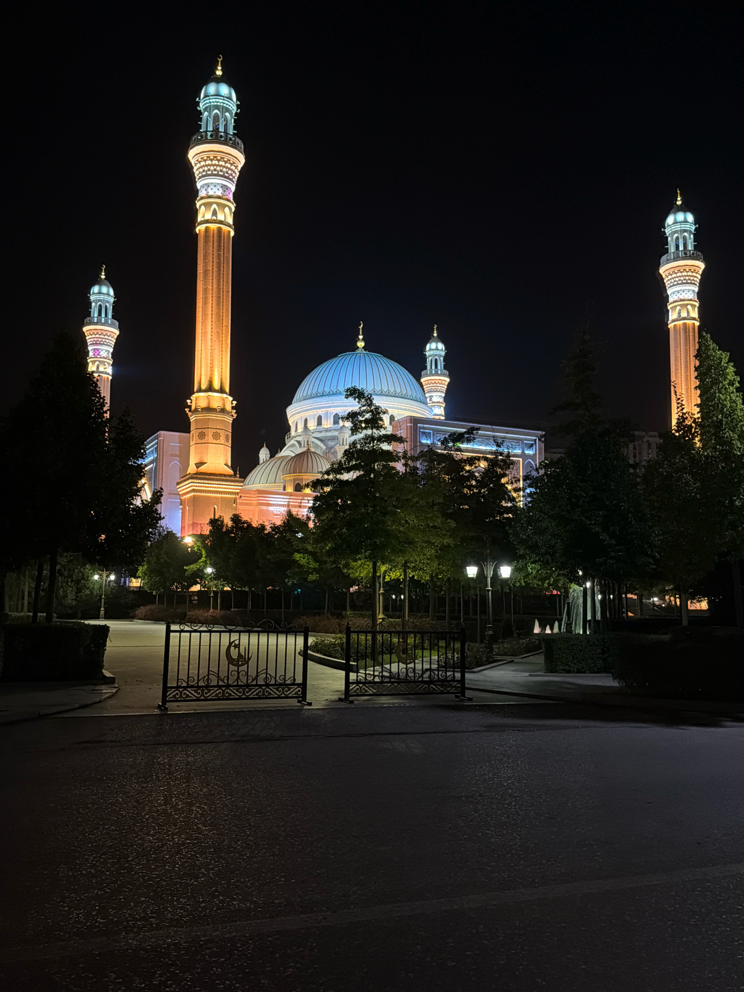 The image depicts the Shali City Mosque in Shali, Russia, at night. The mosque features a prominent central blue dome and four tall, illuminated minarets. The minarets and the main building are lit with warm yellow and white lights, highlighting the architectural details. In the foreground, a black wrought-iron gate with a crescent moon and star emblem stands open, leading to a pathway lined with trees and lampposts. The ground is dark and appears to be asphalt or pavement, with some wet reflections. The overall scene is serene and illuminated by the artificial lighting against the dark night sky. There are no people visible in the image.