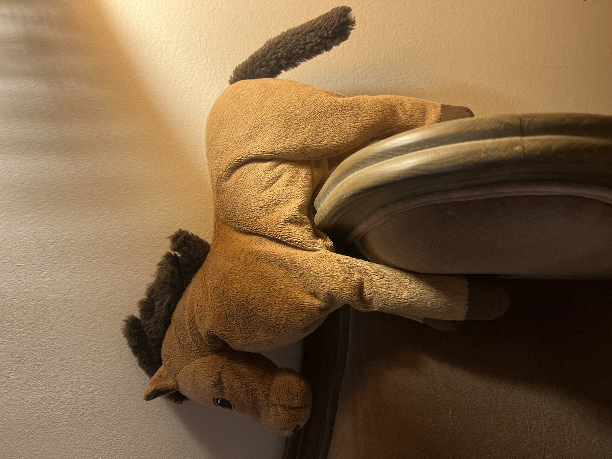 A plush horse toy is resting on the edge of a brown, upholstered piece of furniture. The toy is mostly tan with a brown mane and tail. The image is taken indoors against a light-colored wall. There is no indication of the location being Belgrade, Serbia.