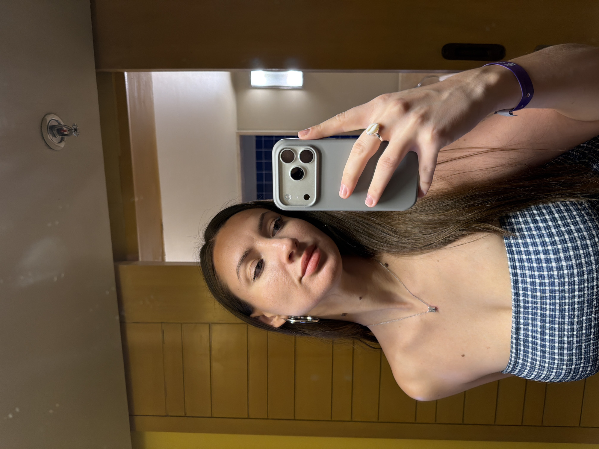 A woman takes a selfie in a bathroom. She is wearing a blue and white checkered strapless top, a silver necklace, hoop earrings, and a ring with a white shell on her right hand. A purple bracelet is visible on her left wrist. The bathroom has wooden paneling on the walls and a large mirror. A fire sprinkler head is visible on the ceiling. The image is taken from a chest-up perspective, showing the woman's upper body and face. The location is identified as a bathroom in Dahab, Egypt.