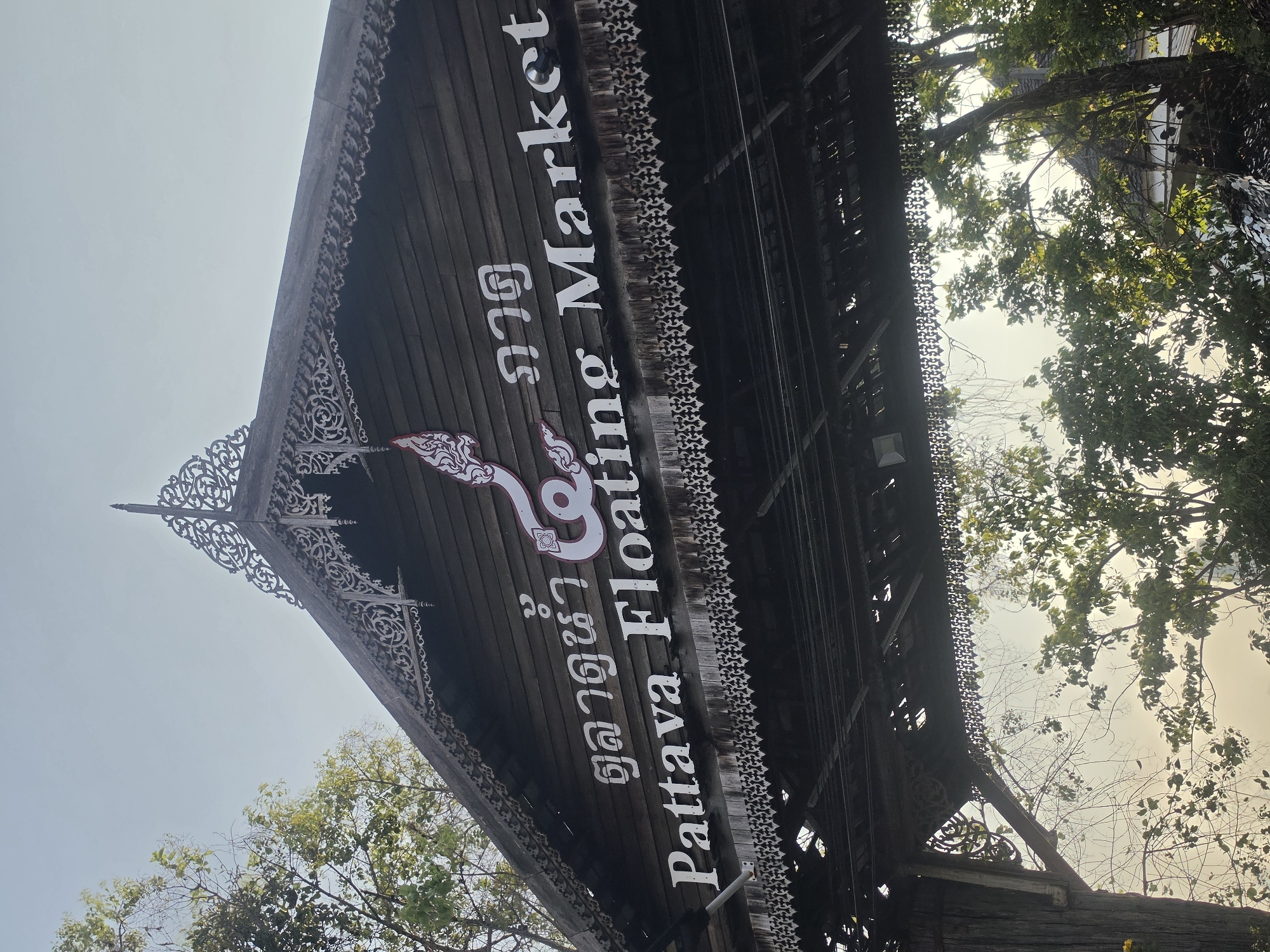 The image shows a low-angle view of the wooden signage for the "Pattaya Floating Market" in Pattaya City, Thailand. The sign is a dark wooden structure with white lettering in both English and Thai. Ornate decorative carvings adorn the edges of the structure. In the background, trees with green leaves are visible against a hazy sky.