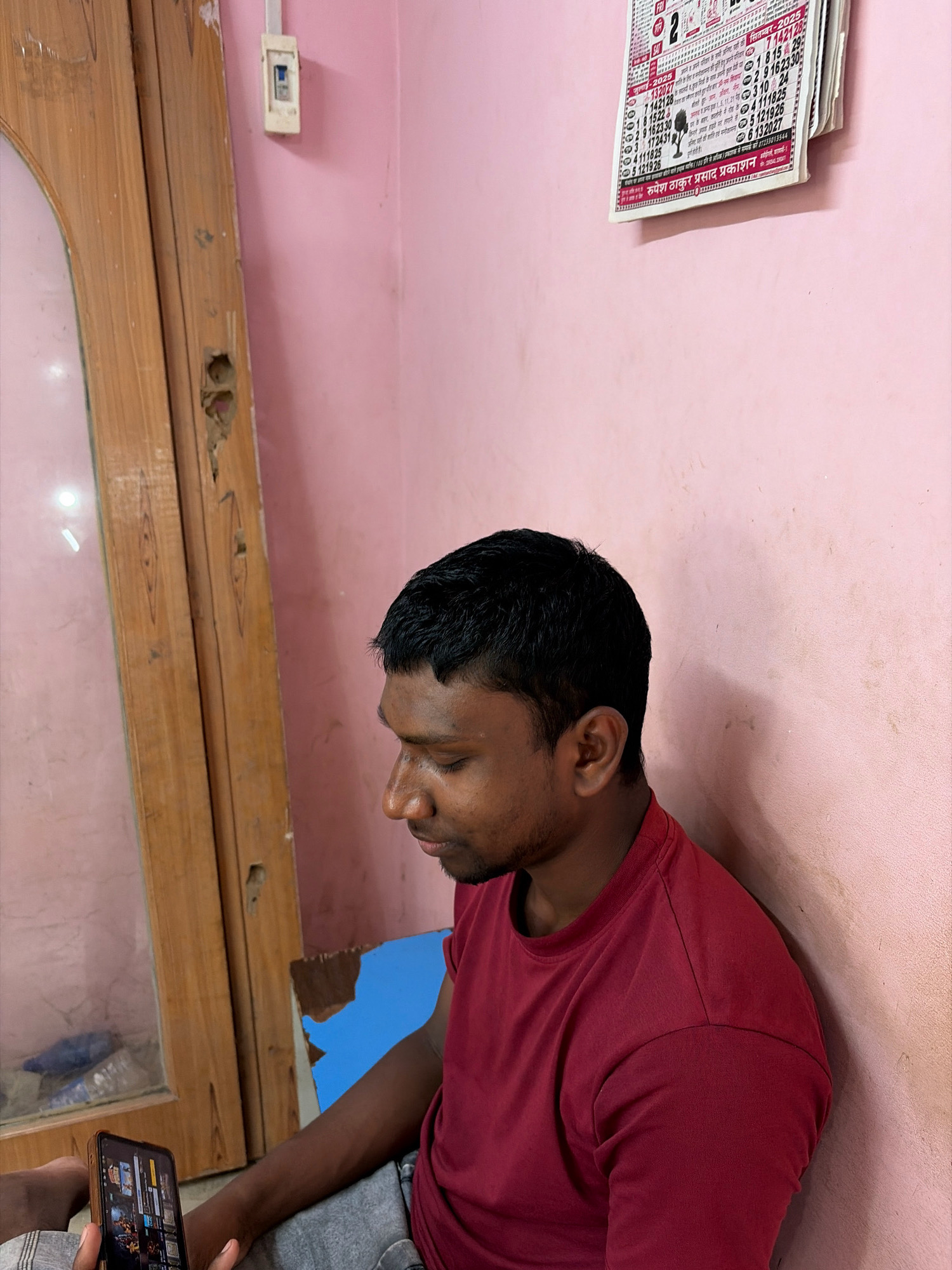 The image depicts an indoor scene, likely a room, with a person holding a smartphone and looking down at it. The person is wearing a maroon t-shirt and gray pants. To the left of the person is a wooden frame, possibly part of a door or window, showing some wear and tear. The wall in the background is painted pink and has a calendar hanging on it. A white electrical switch is also visible on the wall. The overall environment appears simple and possibly a residential setting in Banshtora, India.