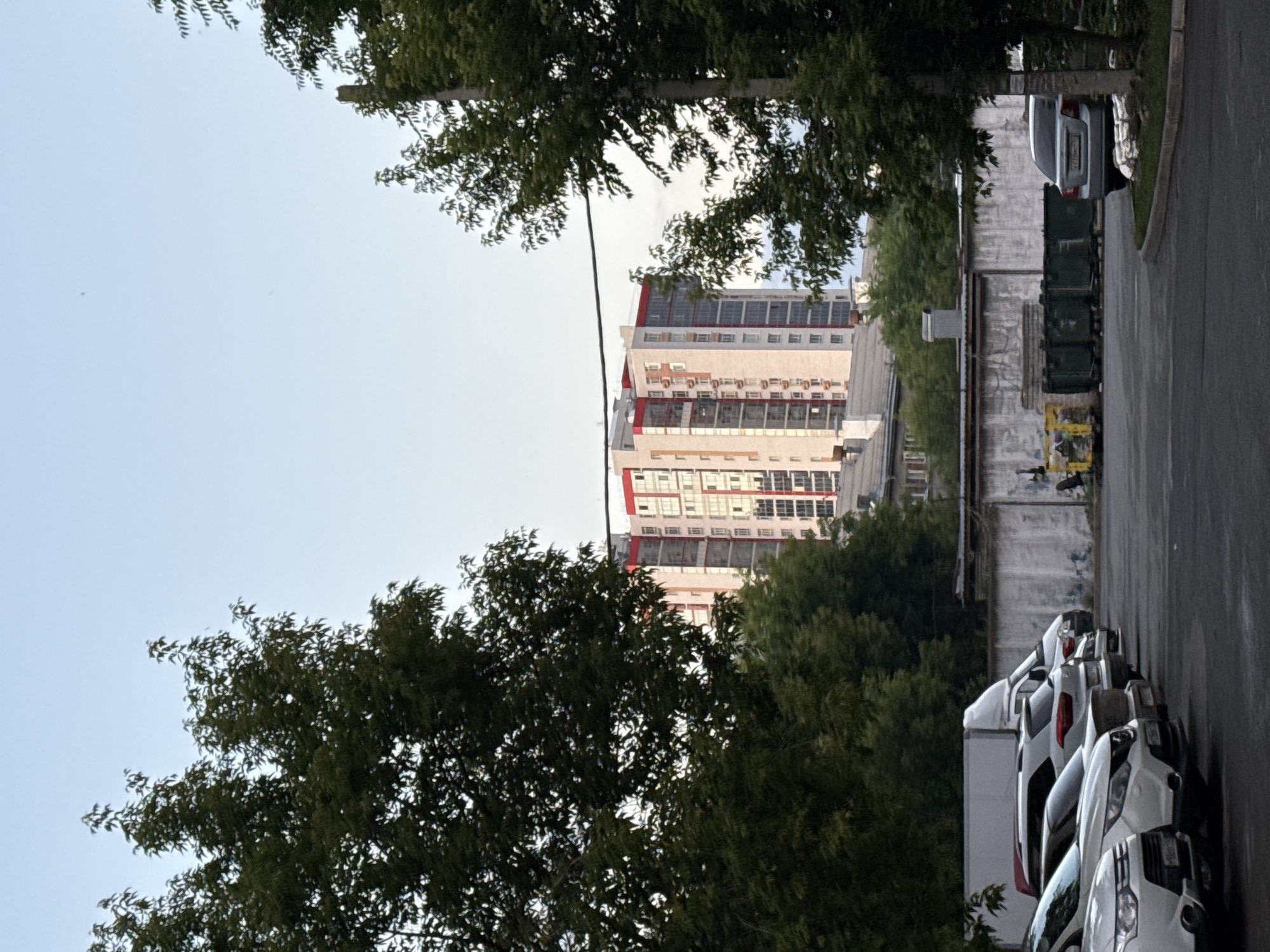 The image depicts an outdoor scene, likely in a residential area of Kazan, Russia, featuring a modern apartment building in the background. The building is tall, with a light-colored facade accented by red trim around windows and the roofline. Several stories are visible, with numerous windows and air conditioning units. In the middle ground, a low concrete wall runs horizontally, behind which are rows of large green trash dumpsters and two smaller yellow containers. Trees with dense green foliage frame the scene from the left and right, partially obscuring the view of the apartment building. In the foreground, a parking lot is visible with several white cars parked in a row. The ground is paved and appears damp. The sky is a clear, pale blue, suggesting either early morning or late afternoon. There are no people visible in the image.