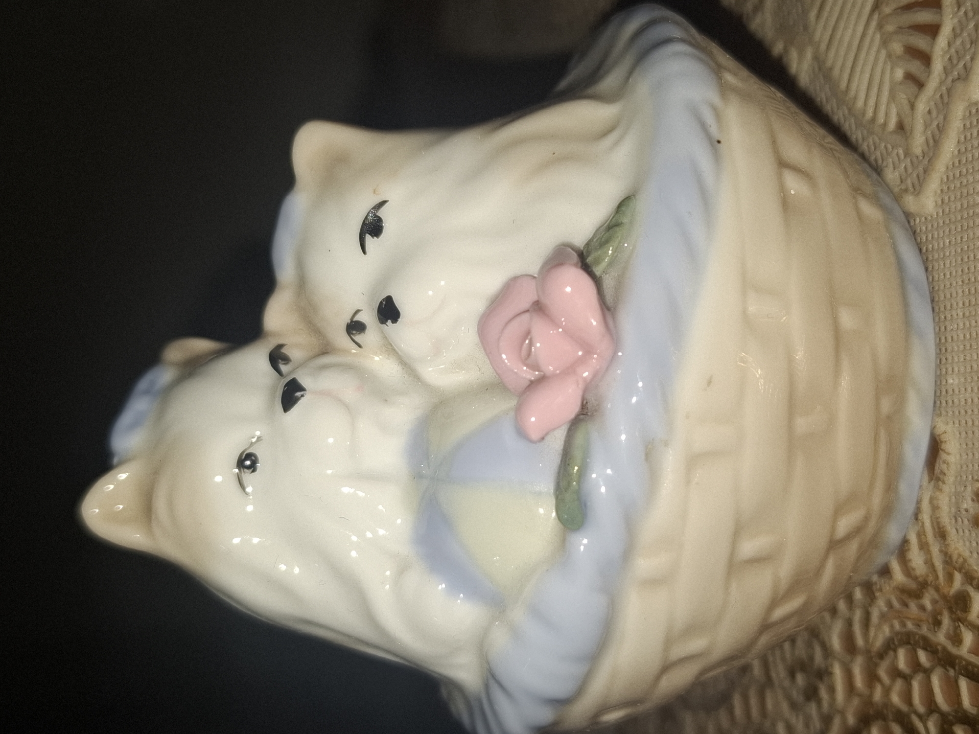 A ceramic figurine of two white kittens nestled in a wicker basket is displayed against a dark, blurred background. The basket has a woven texture and a pale tan color. The kittens are white with black eyes and noses. One kitten is partially obscured by the other, and they appear to be leaning against each other. A pink rose with green leaves is placed in front of the kittens. The basket is lined with light blue material. The figurine is positioned on a textured surface, possibly a tablecloth, with a brown and beige pattern. The overall lighting is dim, with highlights reflecting off the glossy ceramic surface. The scene does not depict any recognizable landmarks or features of Almaty, Kazakhstan.