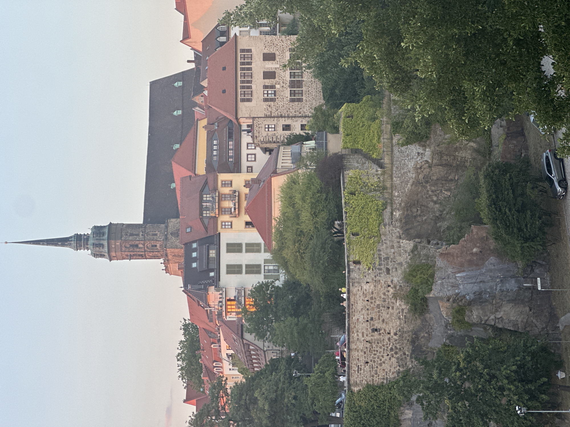The image shows an elevated view of the historic city of Bautzen, Germany, specifically focusing on the Ortenburg Castle and the surrounding cityscape. The foreground features a stone wall covered with ivy, which forms a defensive rampart. Below this wall, a rocky incline is visible, with trees and vegetation growing from it.
In the mid-ground, the Ortenburg Castle complex dominates the scene. Its characteristic stone architecture and red-tiled roofs are clearly visible. A prominent feature is the castle's tall, slender spire, topped with a cross. Numerous other buildings with red and grey roofs are clustered around the castle, creating a dense urban fabric. Some buildings have balconies and windows, suggesting residential or commercial use.
In the background, a clear sky with a hint of pinkish hues, indicating either dawn or dusk, frames the cityscape. Several trees, with lush green foliage, are interspersed throughout the scene, adding natural elements to the urban environment.
In the lower part of the image, a paved area is visible, with a few cars parked on it, including a silver SUV. A lamppost is also present. A small group of people are standing on the stone wall, appearing to be tourists or residents observing the view. Their presence suggests a public area or viewpoint.
The overall scene depicts a picturesque historical setting, with the Ortenburg Castle as the central landmark, overlooking the city and its inhabitants. The lighting suggests a soft, natural light, likely from either the early morning or late evening sun.