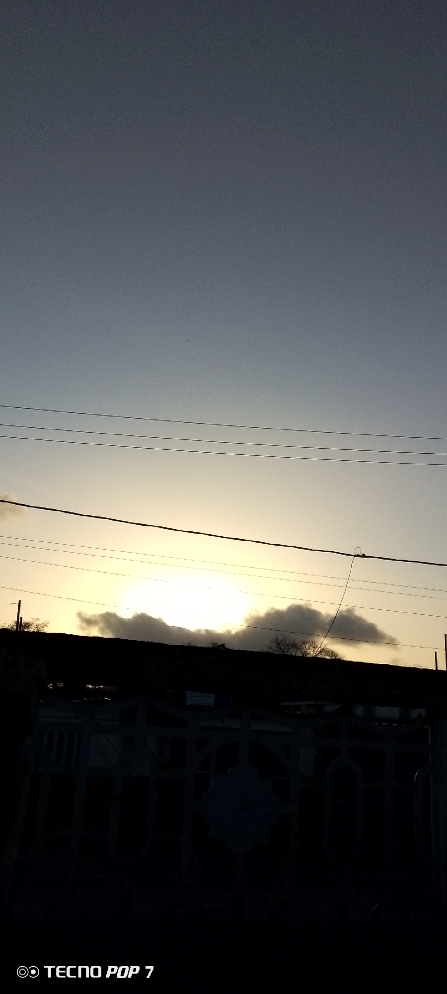 Captured in Altagracia, Venezuela, the image shows a sunset partially obscured by a dark, indeterminate structure and clouds. Overhead power lines are visible against the sky. The foreground features a dark fence. The image was taken with a TECNO POP 7 smartphone, as evidenced by the watermark in the lower-left corner. The scene is shrouded in low light, obscuring details.