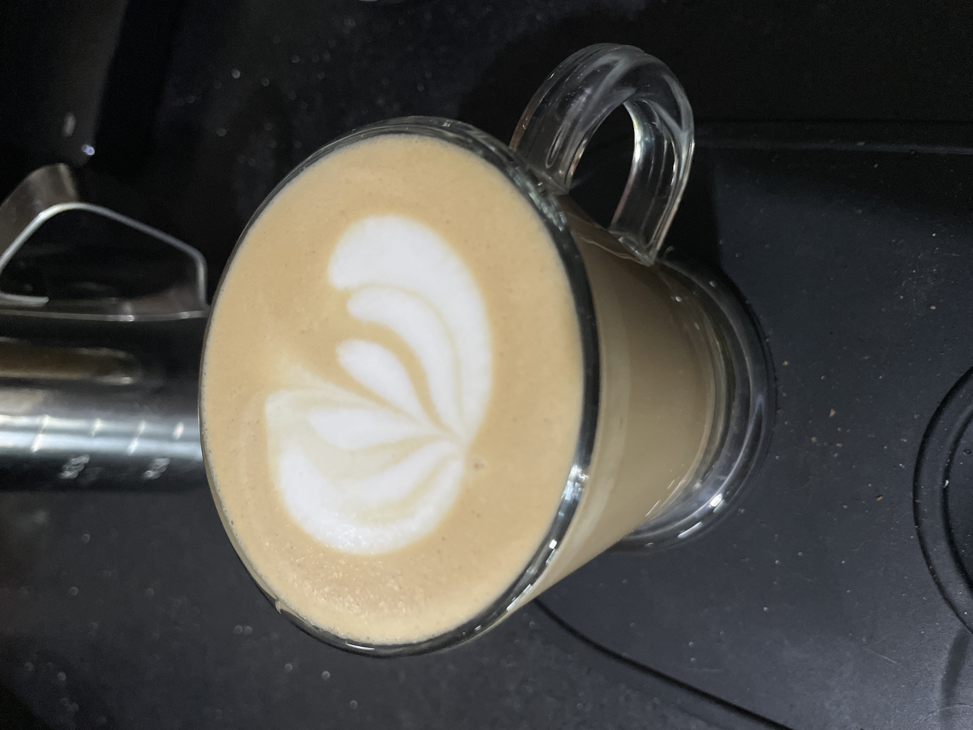 The media file captures a close-up view of a freshly prepared coffee beverage in a clear glass mug. The drink, likely a latte or cappuccino, features a distinct white latte art pattern on its creamy, light brown surface. The intricate design resembles a rosetta or fern, showcasing skilled preparation.
The transparent glass mug has a visible handle on the upper right side. The beverage fills the mug nearly to the brim, highlighting the frothy top. The mug rests on a dark, slightly reflective surface, possibly a kitchen countertop or a coffee machine tray, which exhibits minor specks and reflections. On the far left of the frame, a metallic, curved component of what appears to be a coffee machine or another appliance is partially visible.
The lighting appears artificial, illuminating the coffee and its art. There are no people visible in the image, nor is there any discernible text. The scene conveys an indoor setting, possibly a home kitchen or a cafe in Kuta Selatan, Indonesia. The focus is entirely on the aesthetically pleasing presentation of the beverage.