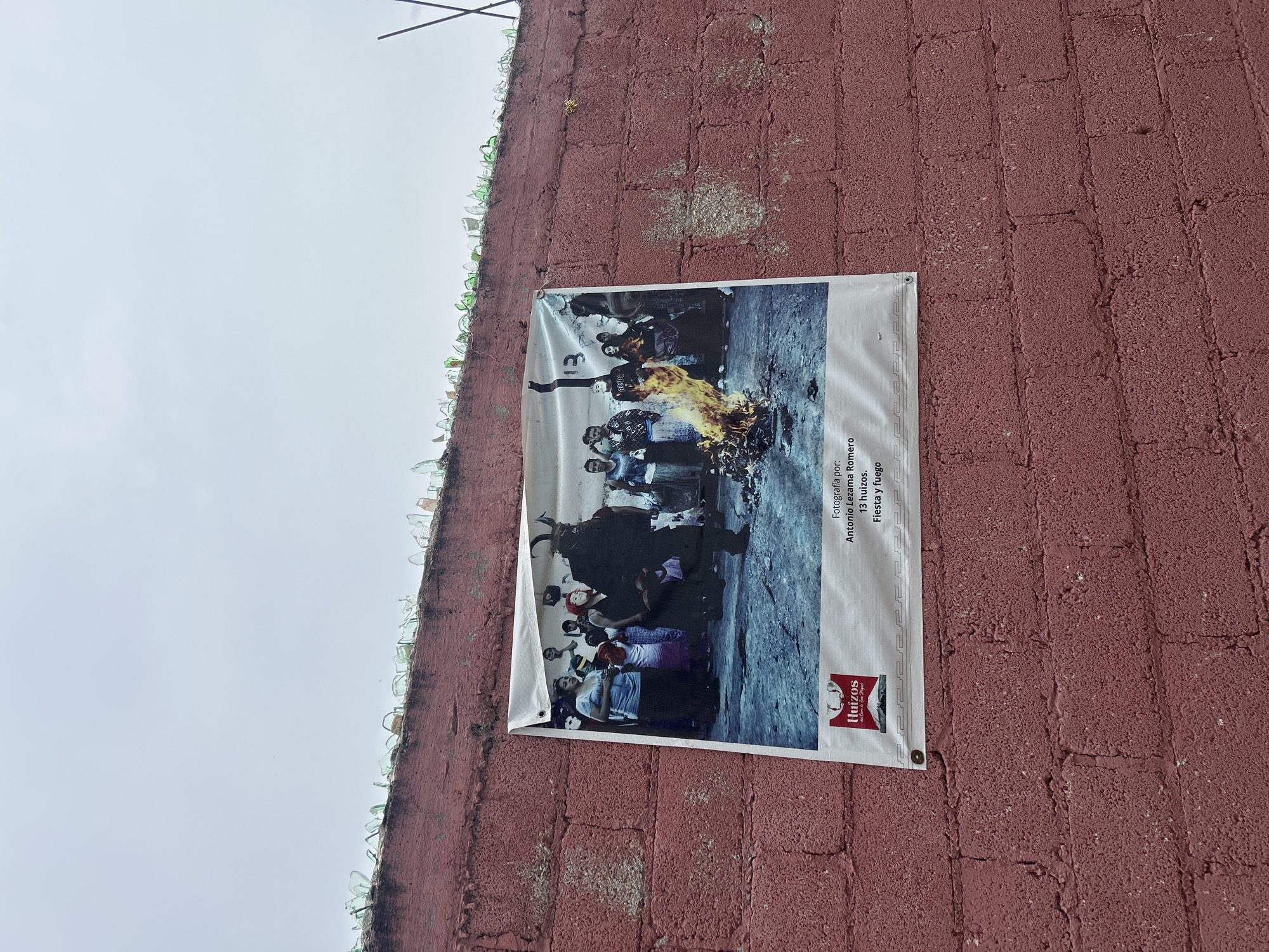 The image shows a poster hanging on a red brick wall. The poster depicts a group of people in what appears to be a ritualistic scene. One person in the center is wearing a dark costume with horns and a mask, and there is a fire burning in front of them. Other people in the poster are wearing masks or are observing the scene. The poster is titled "13 huizos. Fiesta y fuego." The wall is topped with broken green glass bottles, a common security measure. The image was taken in the city of Atlixco, Mexico, likely in the San Miguel neighborhood, as indicated by the logo on the poster which reads "Itzuizos del Corazon de San Miguel." The sky is overcast and the lighting suggests it is daytime.