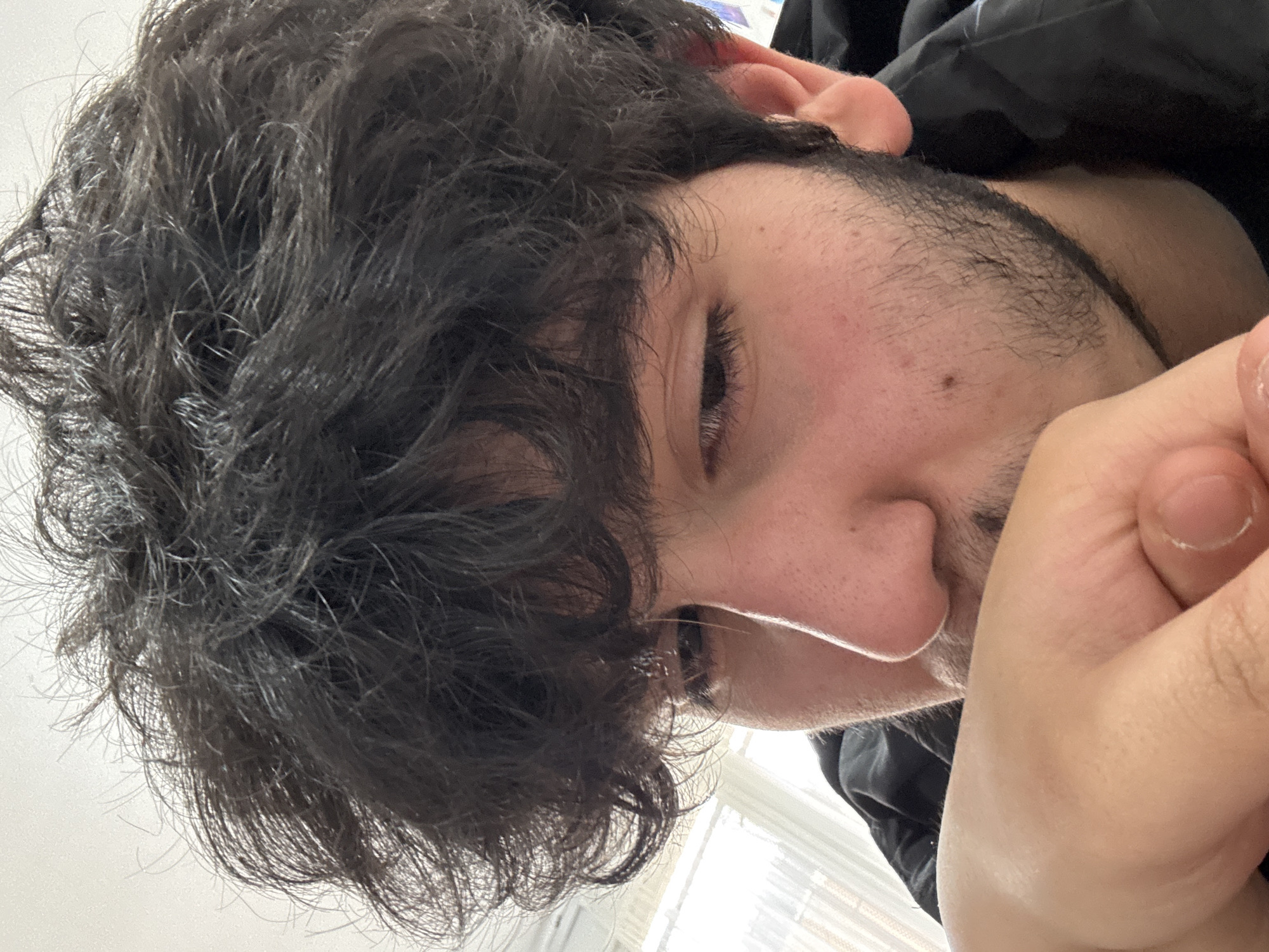 Close-up shot of a person's face and hand. The person has dark, curly hair and is looking down. A black jacket is visible in the background. The person's hand is partially obscuring their face. The location and city of Yerevan, Armenia, are not discernible from the image.