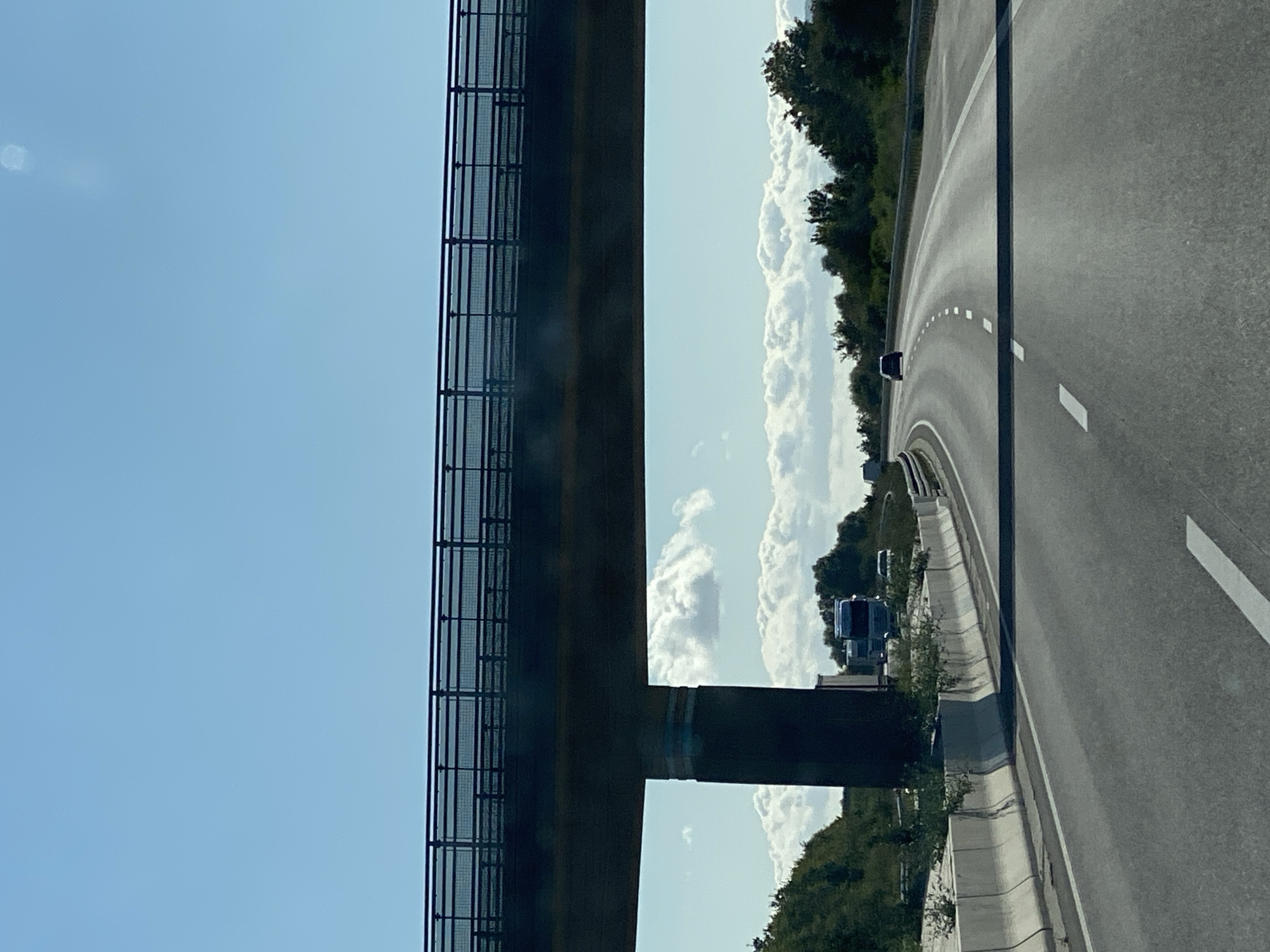 The image depicts a view from a vehicle traveling on a highway, likely an Autobahn in Germany, under a clear blue sky. A large concrete overpass with a metal railing stretches across the upper portion of the frame, supported by a thick concrete pillar on the left. Below the overpass, the highway curves gently to the right. In the distance, a single dark-colored car is visible on the road, and further back, a white truck and other vehicles can be seen. The roadside is lined with concrete barriers and sparse greenery. The overall impression is one of infrastructure within a relatively open, non-urban environment. There is no indication of the specific location within Heringen/Helme, Germany.