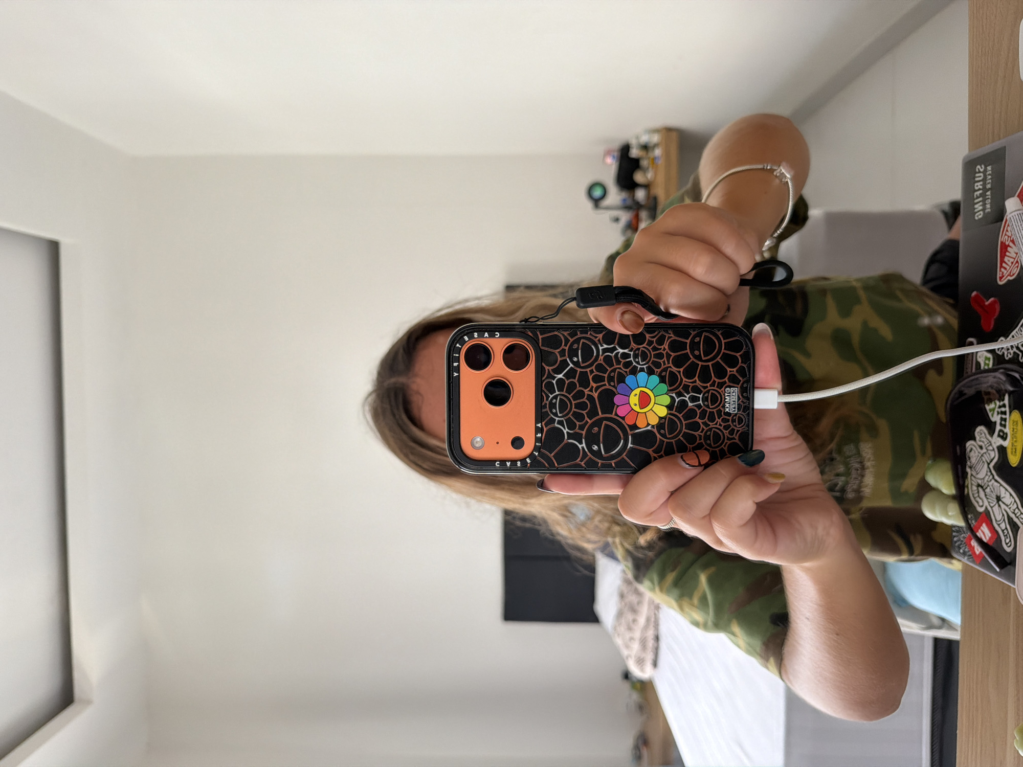 A woman is holding a phone in front of her face, likely taking a selfie. The phone is black with an orange camera area and a design featuring black flowers with a multicolored center flower on the back. It is connected to a white charging cable. She is wearing a camouflage print shirt and a silver bracelet on her left wrist. Part of a table with various items on it is visible on the right side of the frame, including a laptop with stickers, a green glass item, and other objects. A shelf with items on it is in the background. The walls are white. The location is presumed to be indoors in Kuta Selatan, Indonesia.