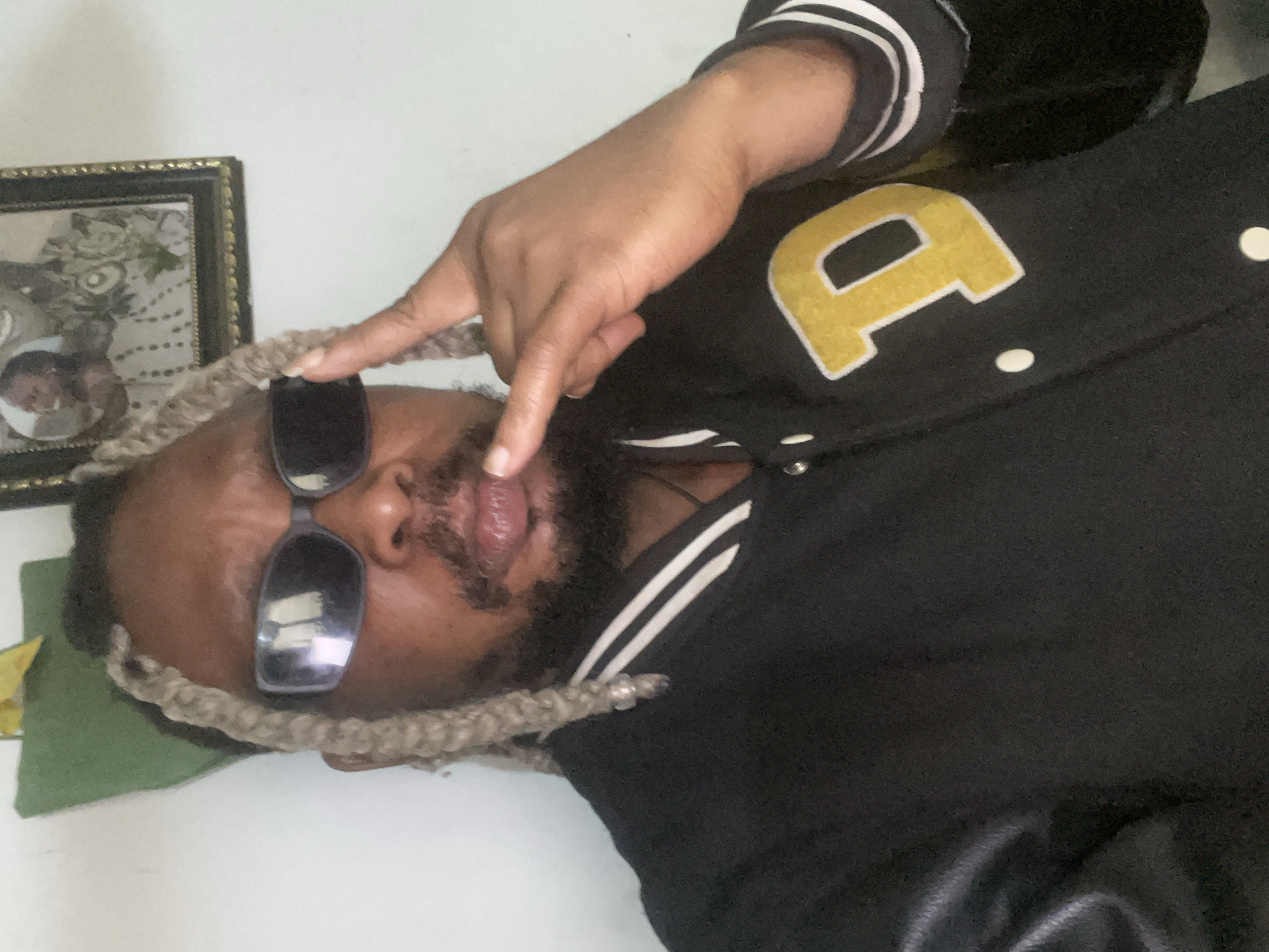 A medium shot captures a Black man indoors, facing the camera. He has light-colored dreadlocks, a beard, and is wearing dark sunglasses. He gestures with his right hand, index and pinky fingers extended, near his forehead and sunglasses. He is dressed in a black varsity jacket with white stripes on the collar and a large yellow "D" emblem on the left chest. The background features a white wall with a framed picture and a green object hanging on the wall. The lighting suggests an indoor setting with possibly overhead light. The scene does not contain any specific landmarks that would allow for a precise location within Jos, Nigeria.