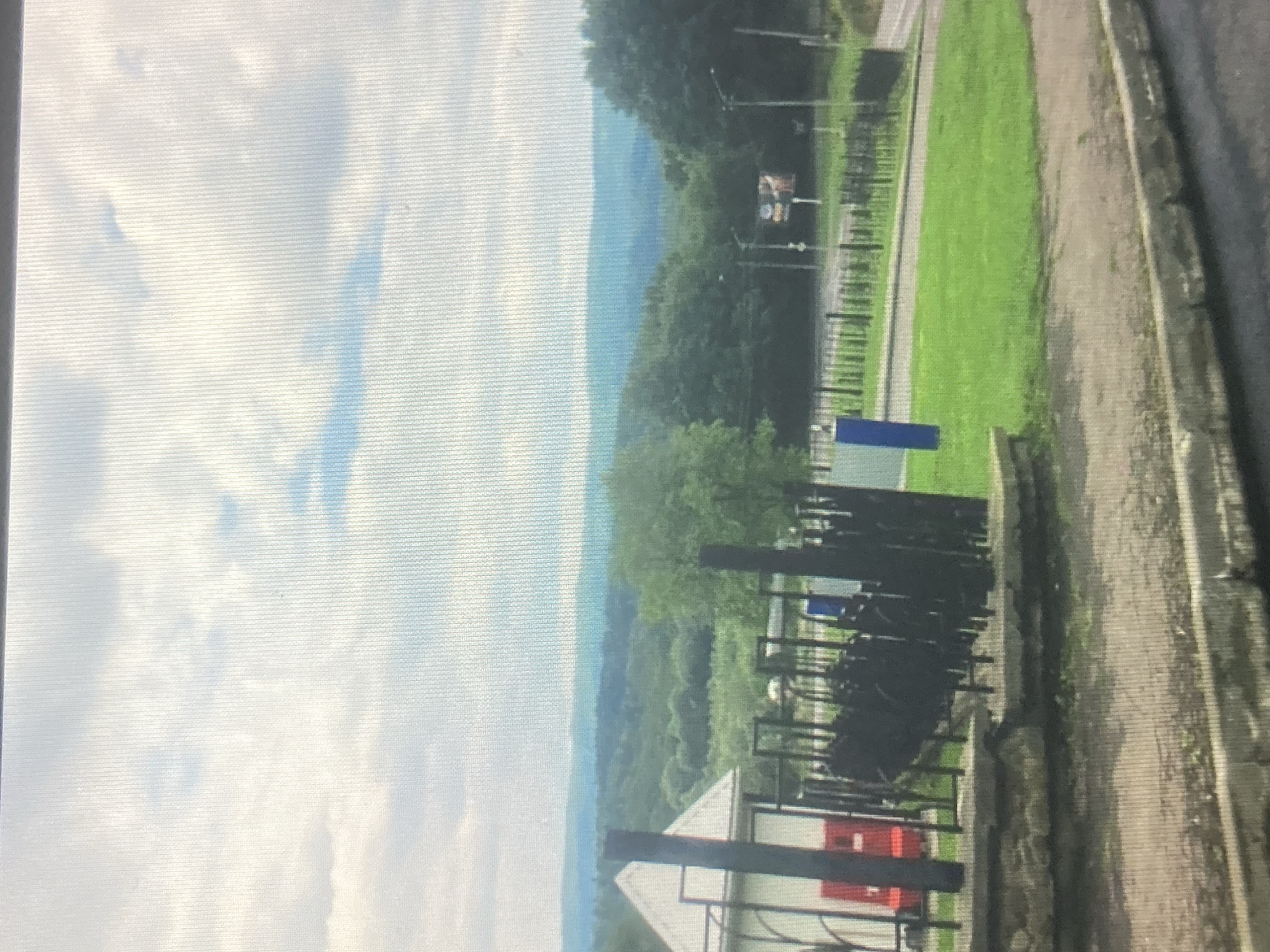 The image depicts a view of a landscape with rolling hills and trees under a cloudy sky. In the foreground, there is a white building with a triangular roof, partially obscured by black metal fencing. To the left of the building, a red object is visible. A green grassy area with a pathway runs through it, leading towards a road in the mid-ground. Several streetlights are present along the road, and a billboard with an image is visible in the distance. The overall scene suggests an outdoor environment, possibly a park or a scenic overlook, with some infrastructure present. There are no people visible in the image. The location is not identifiable as the city of Dnipro, Ukraine, based on the visual information provided.