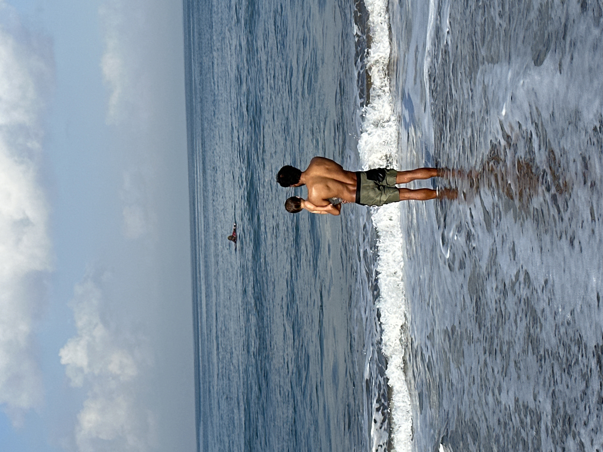 A man and a baby are standing in the ocean off the coast of North Kuta, Indonesia. The man is holding the baby in his arms, and they are both looking out at the water. In the distance, there is a surfer on a surfboard. The sky is blue with a few clouds. The waves are breaking on the shore, and the water is clear and blue.