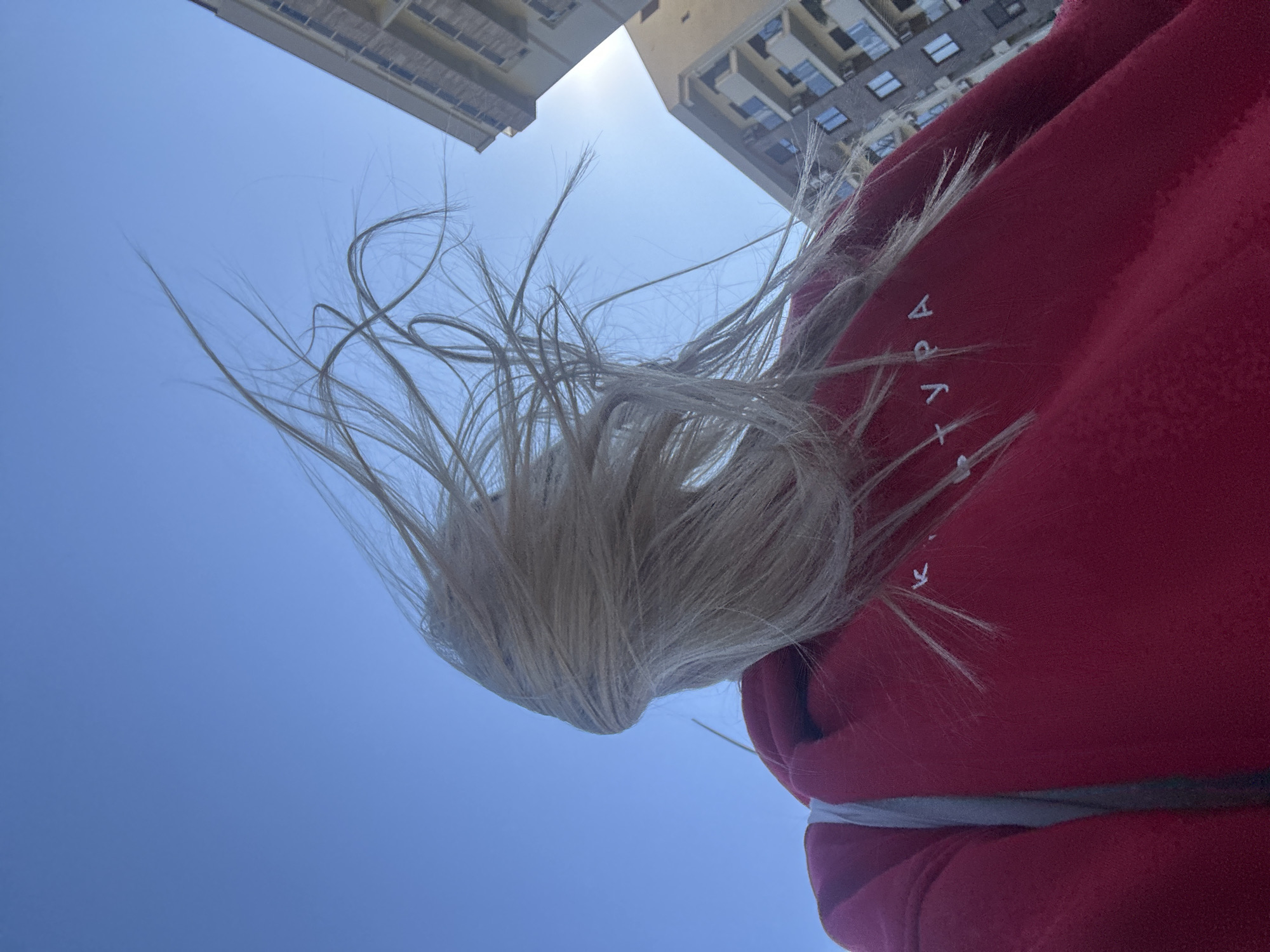 The image is a low-angle shot looking up towards a clear blue sky. Two tall buildings are visible in the upper portion of the frame, suggesting an urban environment. The focus of the image is a person wearing a red hoodie with white lettering. The person's blond hair is blowing upwards, obscuring their face. Based on the architecture, urban landscape, and light, the location is possibly Budva, Montenegro.