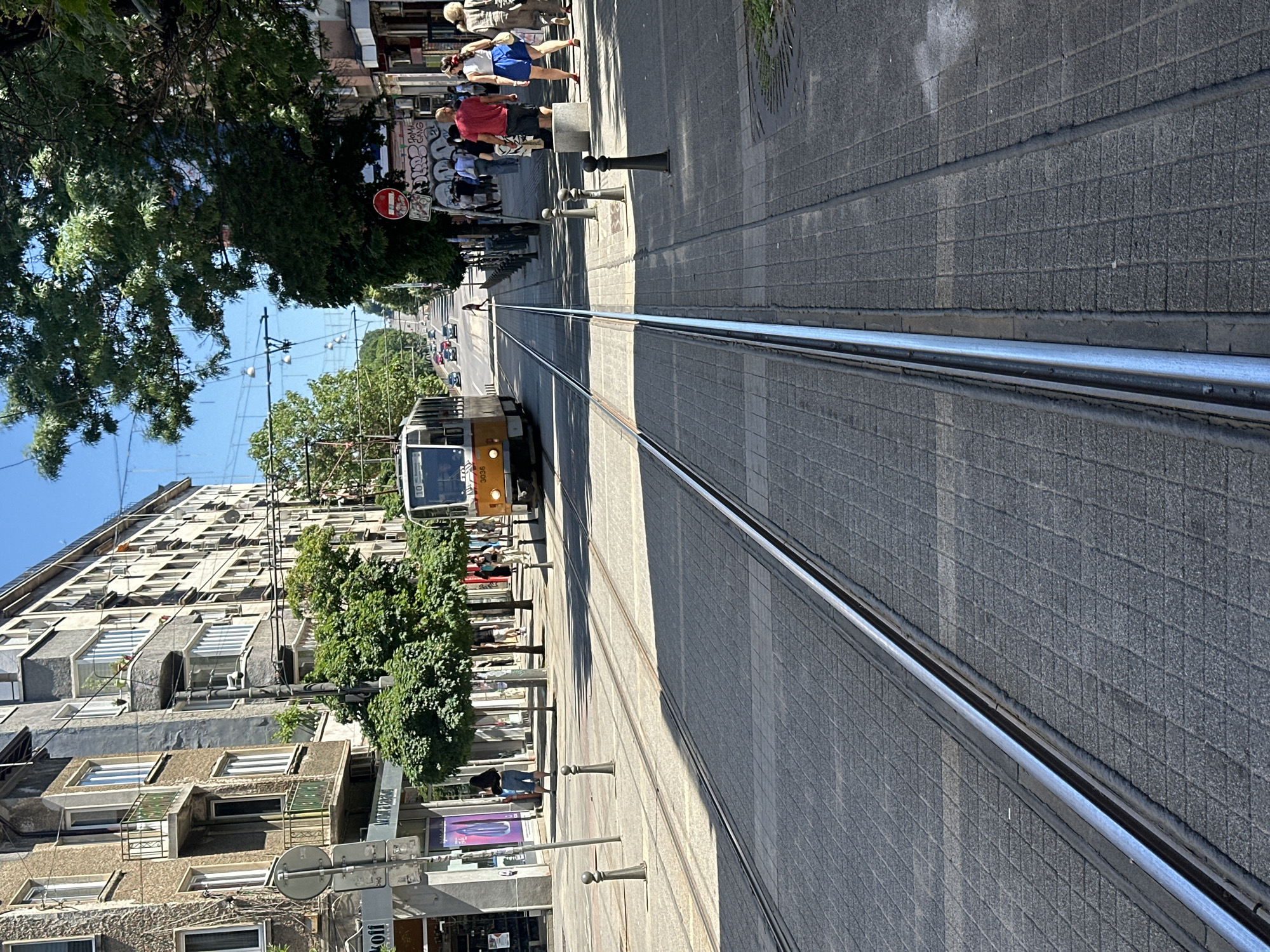 An eye-level medium outdoor shot captures a street scene in an urban environment. The focal point is a tram track running down the center of the paved street, with a yellow and white tram moving away from the viewer in the distance. On the right side of the street, a sidewalk is lined with trees, lampposts, and a few pedestrians. Buildings flank the street, some appearing older and weathered, others more modern. The sky is clear and blue, indicating a sunny day. The overall impression is that of a busy city street during daylight hours.