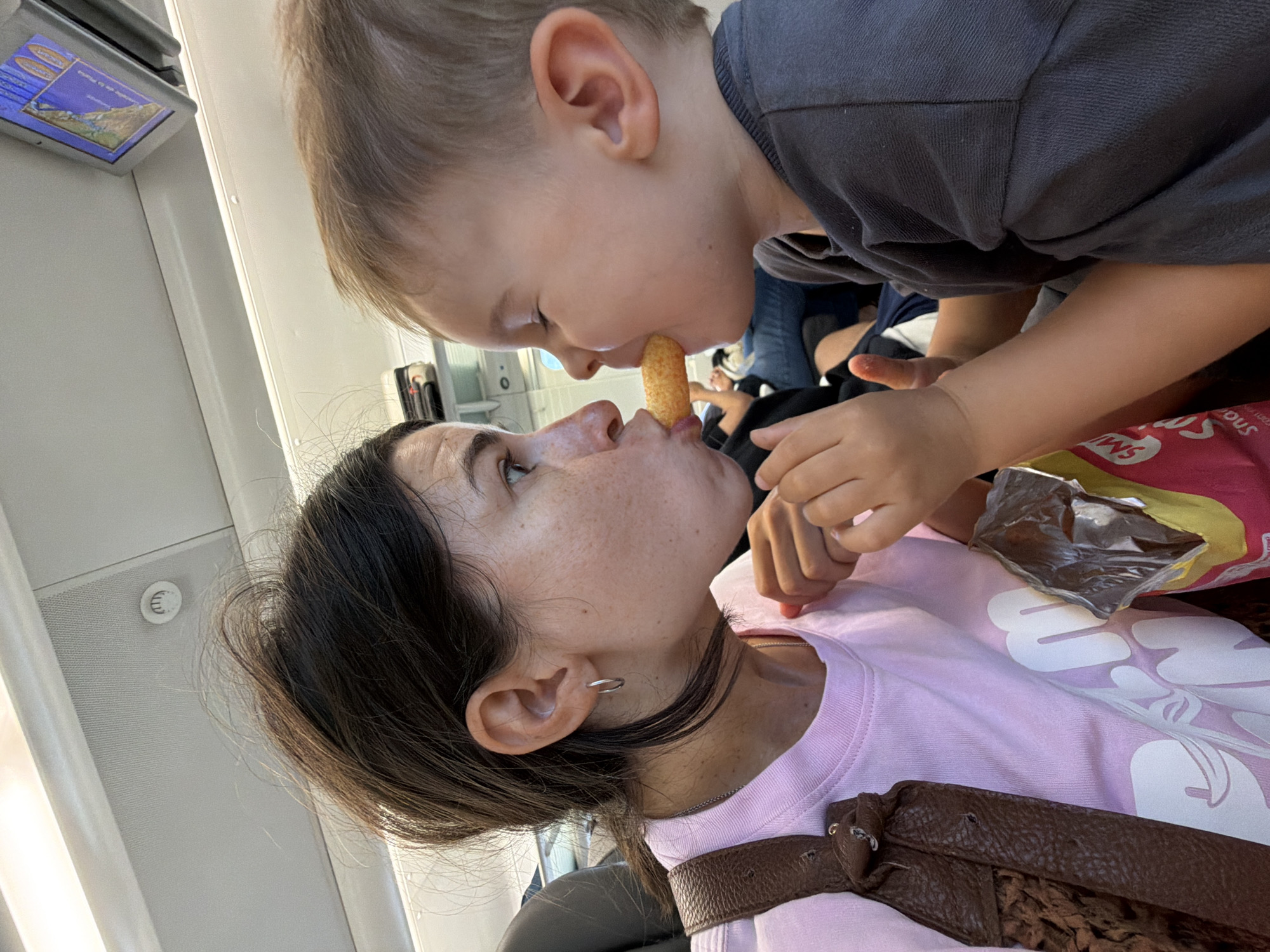 A woman and a young child are on what appears to be public transportation, possibly a train or airplane, with overhead compartments and seating visible. They are positioned close together, facing each other. The woman, wearing a pink t-shirt and a brown shoulder bag, is looking at the child. The child, wearing a grey shirt, is reaching out a snack to the woman, who is also reaching out to take it. Their faces are very close, with the snack between their mouths. A bag of snacks is visible in the foreground. The overall scene captures a moment of playful interaction between the two. The location is identified as Tarragona, Spain.