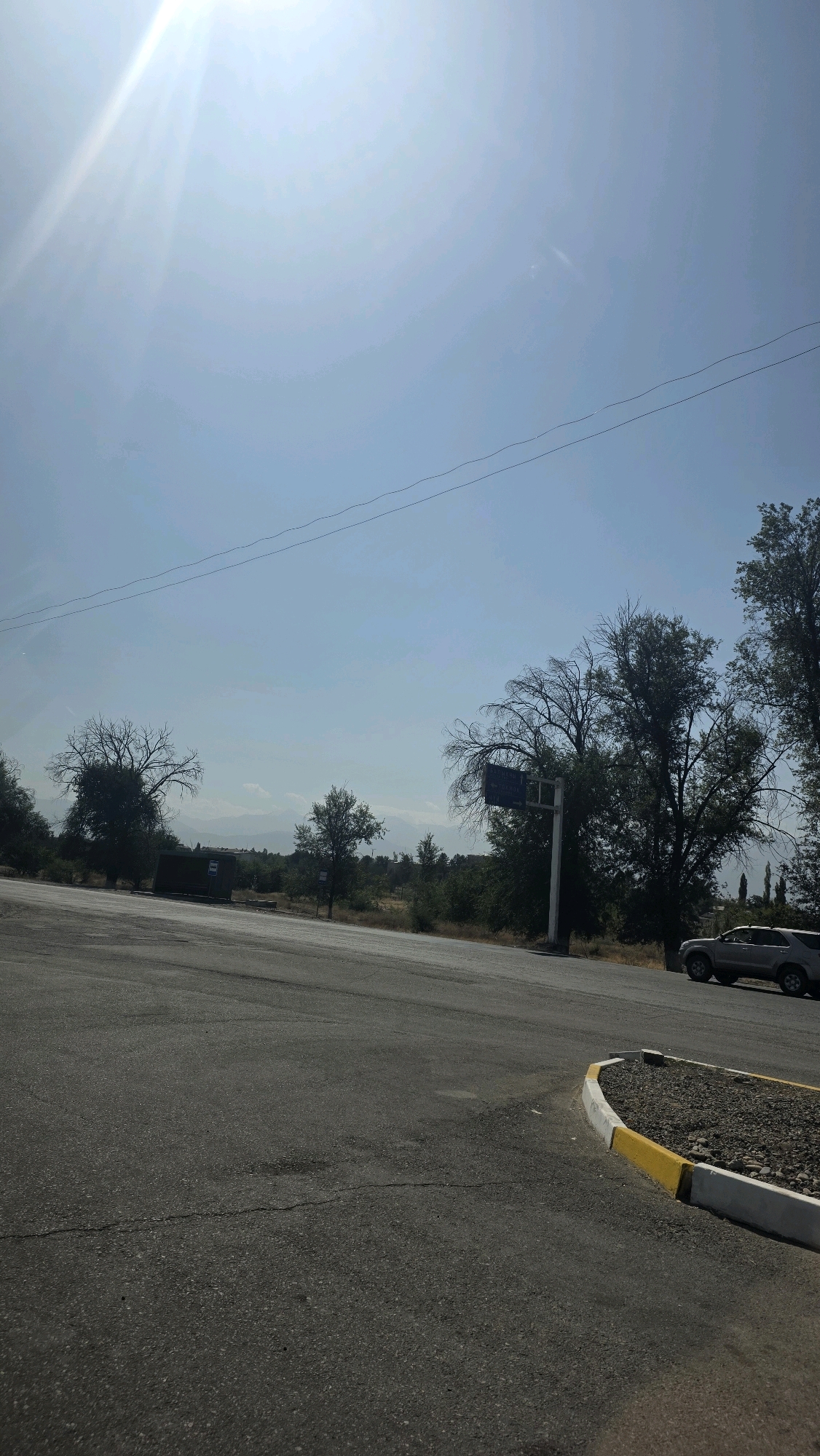 The image shows a section of a road in Tokmok City, Kyrgyzstan, under a clear blue sky with strong sunlight casting bright glare and lens flares. The road is paved with asphalt, showing some cracks and wear. In the foreground, the road curves to the right, with a raised curb that is painted with yellow and white stripes. Beyond the road, there is a dry, grassy area with scattered trees, some bare and others with foliage. In the middle ground, a bus stop shelter is visible, along with a utility pole and a blue street sign. Further in the distance, mountains are faintly discernible against the horizon. A silver SUV is parked on the right side of the road. Power lines are strung across the sky.