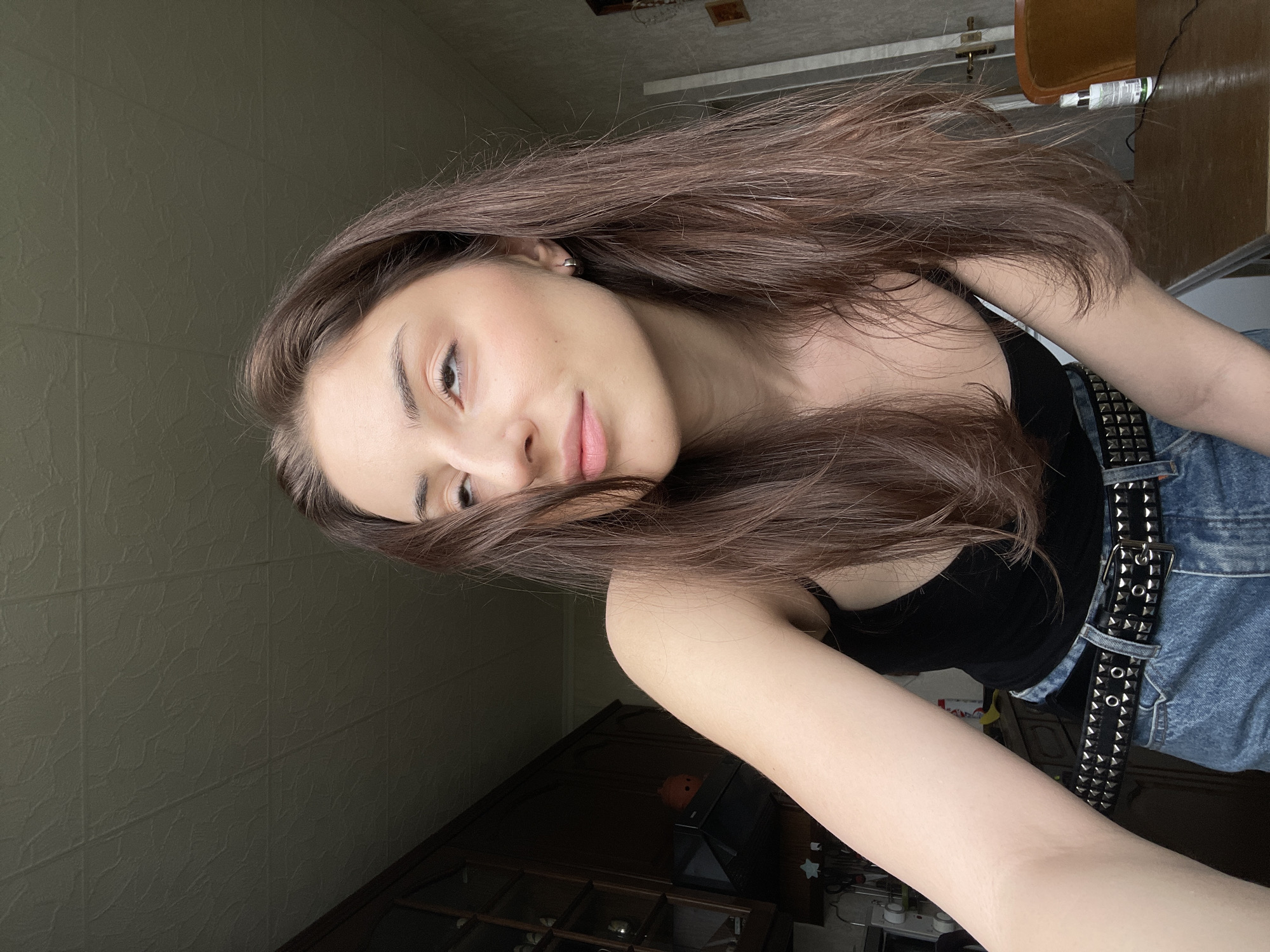 A young woman with long, dark brown hair is captured in a selfie from the chest up. She is wearing a black tank top and blue jeans, accessorized with a studded black belt. Her head is tilted, and her gaze is directed upwards and to the right, away from the camera. The background appears to be an interior space, possibly a kitchen or dining area, with wooden cabinets visible on the left. A table with a chair can be seen on the right. The lighting suggests it might be daytime, with light filtering in from the right side of the frame. There are no discernible elements that place this image specifically in Moscow, Russia, or any other particular city.
