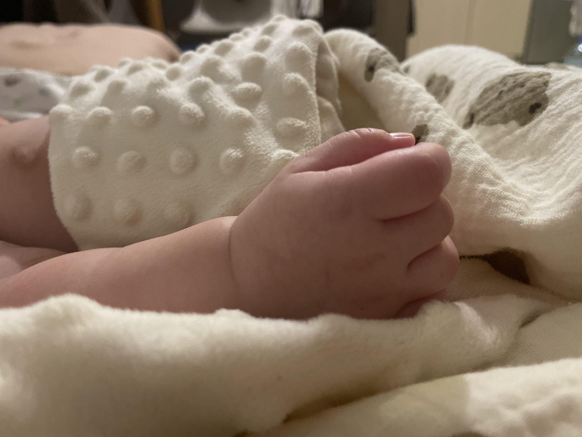 A close-up photograph shows a baby's arm and hand resting on several soft blankets. One blanket is white with raised, textured circles, while another is off-white with a faint animal print. The baby's hand is clenched in a loose fist. The background is blurry, showing only indistinct shapes and colors, suggesting the photo was taken indoors. The image does not contain information related to Bar, Montenegro.