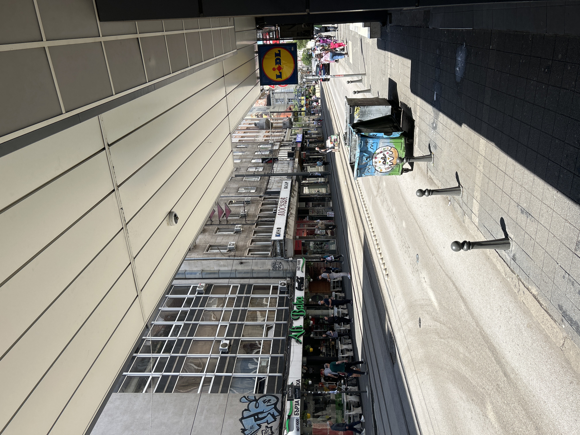 This image captures a street view in an urban setting. Dominating the scene on the left is a tall, modern building with a patterned facade, possibly made of panels or large windows. Opposite this building, across the street, are older, multi-story buildings displaying various advertisements and storefronts at street level. A Lidl supermarket sign is visible farther down the street.
The street itself is paved and equipped with bollards lining the edge, separating pedestrian traffic from the road. There are people present on the sidewalks, some walking, some appearing to be seated outside the storefronts. A pair of trash bins, decorated with graffiti, are also positioned along the sidewalk. The angle of the shot, combined with the high perspective, offers a compressed view of the street, emphasizing the depth of the urban landscape.