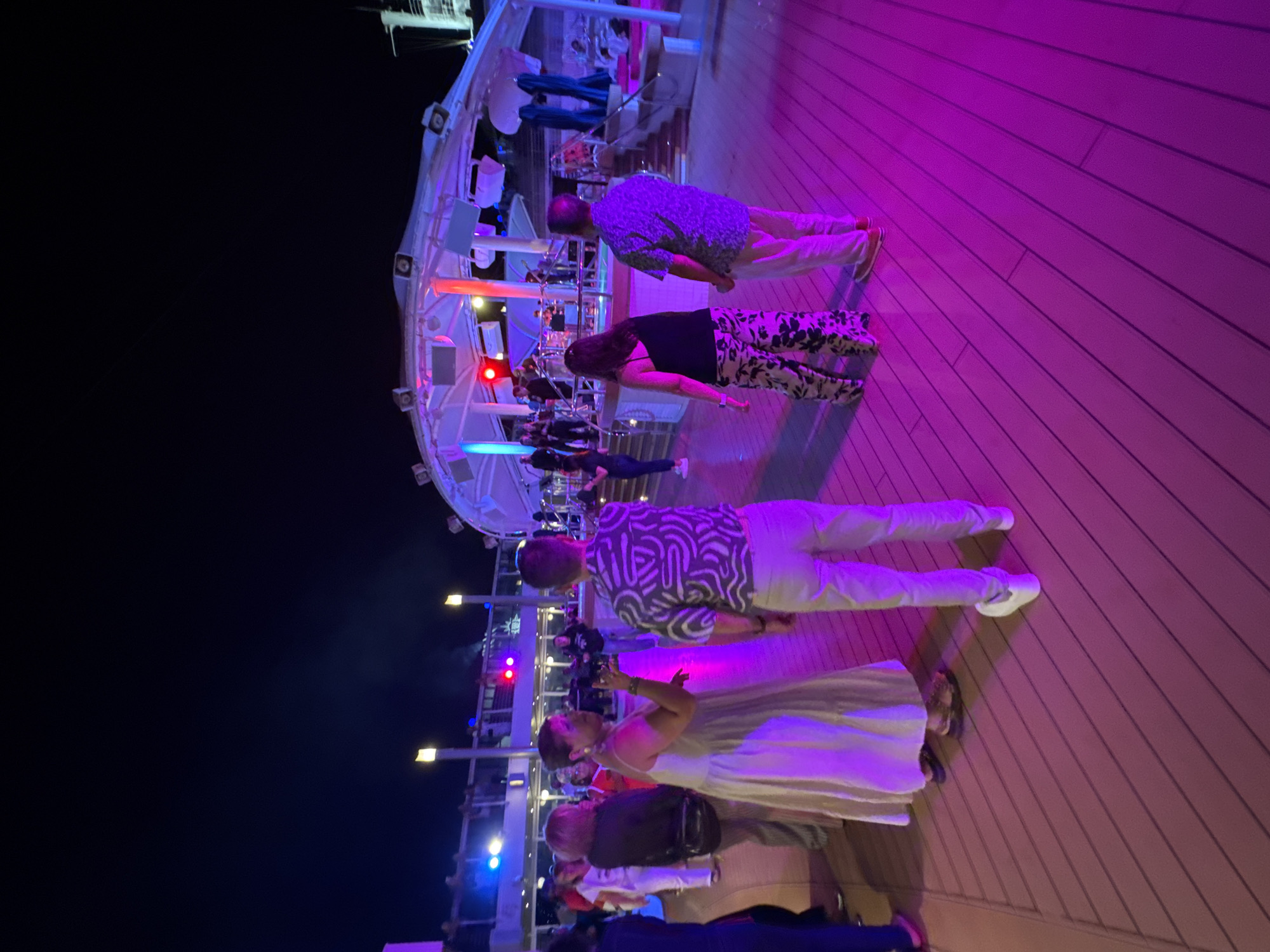 The image depicts a nighttime scene on what appears to be a ship deck or a large outdoor venue with a wooden floor bathed in pink and purple light. Several individuals are present, some standing and others seemingly dancing or conversing. In the background, a stage area is visible with a band performing and lights illuminating the structure. The atmosphere suggests a party or entertainment event. The location is not the city of Ibiza, Spain, but rather appears to be a cruise ship or a resort at night.