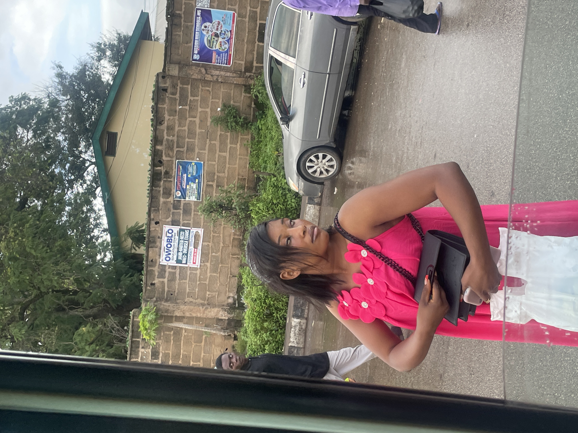 In Barakin Lerawi, Nigeria, a young woman with dark hair, wearing a bright pink dress adorned with floral appliqués, stands with a black clutch bag in her hands. She is looking to her right with a sideways glance. Behind her, a man in a black shirt and light gray pants is walking away, his head turned back towards her with a startled expression. The scene takes place outdoors, on a wet asphalt road, suggesting it has recently rained. In the background, a brick wall is visible, with several posters attached to it. One poster reads "OWOBLO EDUCATION CENTRE" and advertises various exams. Another poster displays "MISSION HIGH SCHOOL, JOS". A light-colored sedan is parked on the right side of the road. A building with a green roof is visible behind the wall. The overall environment appears to be an urban setting with some greenery.