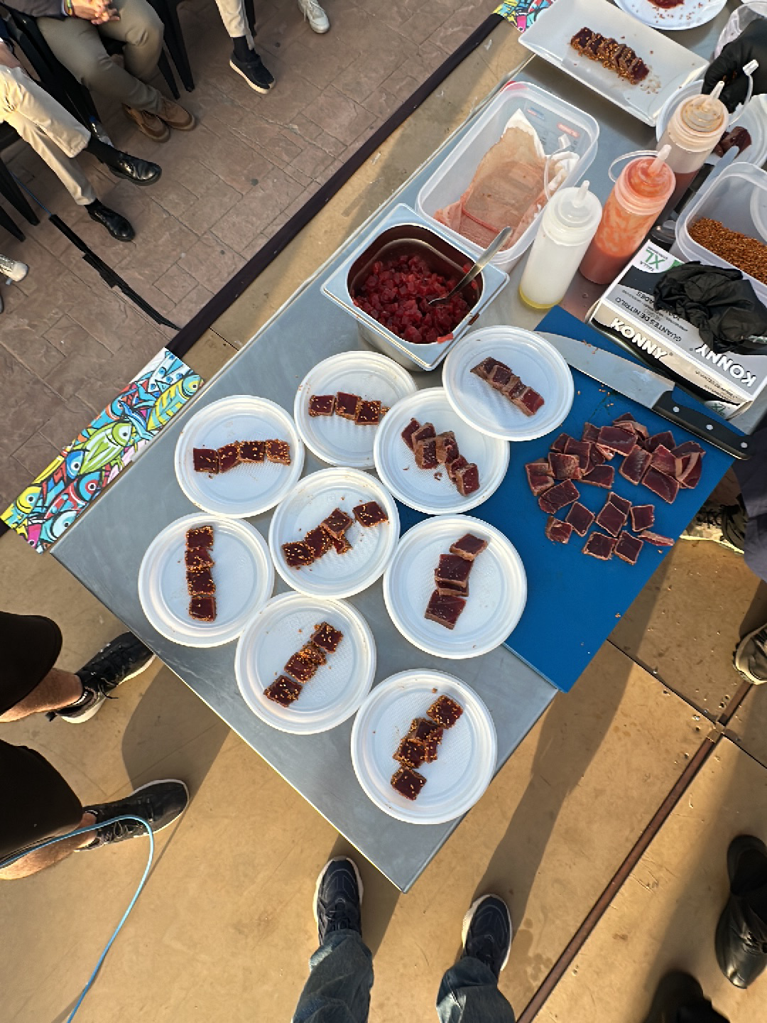 The image shows an outdoor food preparation setup, likely a culinary demonstration or tasting event, possibly in Barbate, Spain. A stainless steel table is the central feature, upon which are several white plastic plates arranged with portions of what appears to be seared tuna, each topped with sesame seeds. Additional ingredients are present in containers, including diced raw tuna, sauces in squeeze bottles, and possibly spices. A blue cutting board holds larger pieces of tuna, alongside a large knife. A box of gloves is visible on the table. Around the table, several people are standing, their legs and feet visible in the frame's edges. The environment has a sunlit, outdoor feel with a patterned floor.