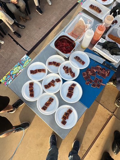 The image shows an outdoor food preparation setup, likely a culinary demonstration or tasting event, possibly in Barbate, Spain. A stainless steel table is the central feature, upon which are several white plastic plates arranged with portions of what appears to be seared tuna, each topped with sesame seeds. Additional ingredients are present in containers, including diced raw tuna, sauces in squeeze bottles, and possibly spices. A blue cutting board holds larger pieces of tuna, alongside a large knife. A box of gloves is visible on the table. Around the table, several people are standing, their legs and feet visible in the frame's edges. The environment has a sunlit, outdoor feel with a patterned floor.