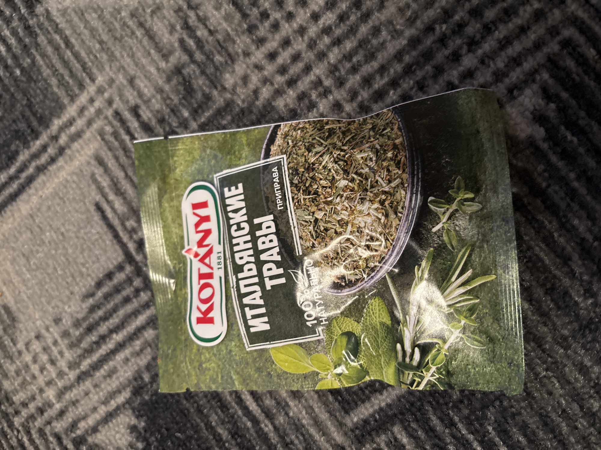 The image shows a package of "KOTANYI ITALIAN HERBS" seasoning. The package is green with red lettering and features a bowl filled with dried herbs. Fresh herbs, possibly sage and rosemary, are also depicted on the packaging. The product is placed on a dark, textured surface, possibly a carpet or fabric. The location is identified as Chernogorsk, Russia.