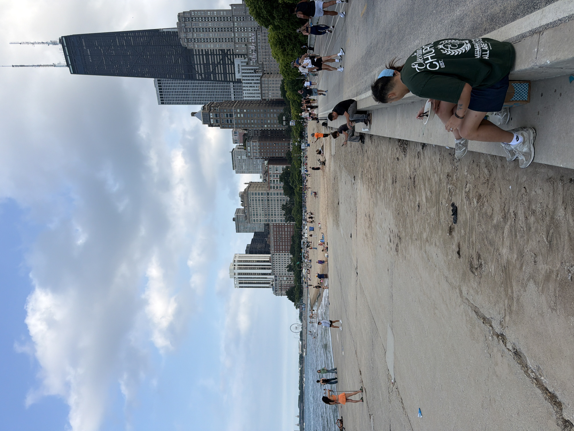 The image captures a daytime scene at a beach in Chicago, Illinois, with the iconic John Hancock Center visible in the background. The foreground shows a concrete path bordering a sandy beach. On the path, a man and a woman are seated, with the man eating from a bowl. Several other people are scattered across the beach and the path, some walking, others sitting or playing. The water of Lake Michigan is visible to the left. The sky is partly cloudy.