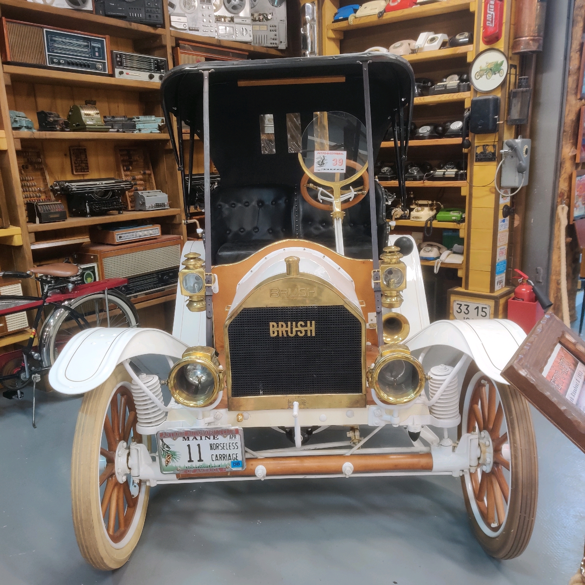 An antique white Brush car with brass accents and wooden wheels is parked indoors, likely in a museum or collection. The car has a black fabric top and leather seats. The front grille bears the "BRUSH" name in gold lettering. A license plate from Maine, reading "11 HORSELESS CARRIAGE," is visible on the front. The background features shelves filled with vintage electronics such as radios, typewriters, calculators, and telephones, suggesting a retro or historical theme. The lighting is artificial. There are no people present in the image. The scene is static and presented from a frontal, slightly low-angle perspective.
