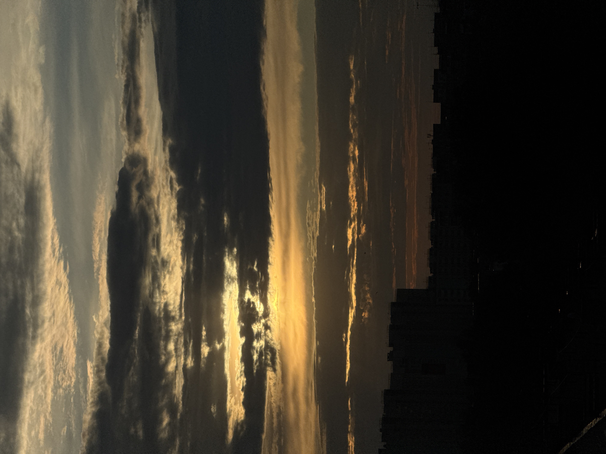 The image shows a sunset sky above what appears to be the skyline of Nizhny Novgorod, Russia. The sky is filled with layered clouds, ranging in color from light grey to dark grey and black, with streaks of golden light piercing through the formations. The silhouette of a building or structure is visible against the dark part of the sky on the right, adding a faint outline of urban landscape. The scene conveys a sense of twilight or dusk, with the interplay of light and shadow creating a dramatic effect.