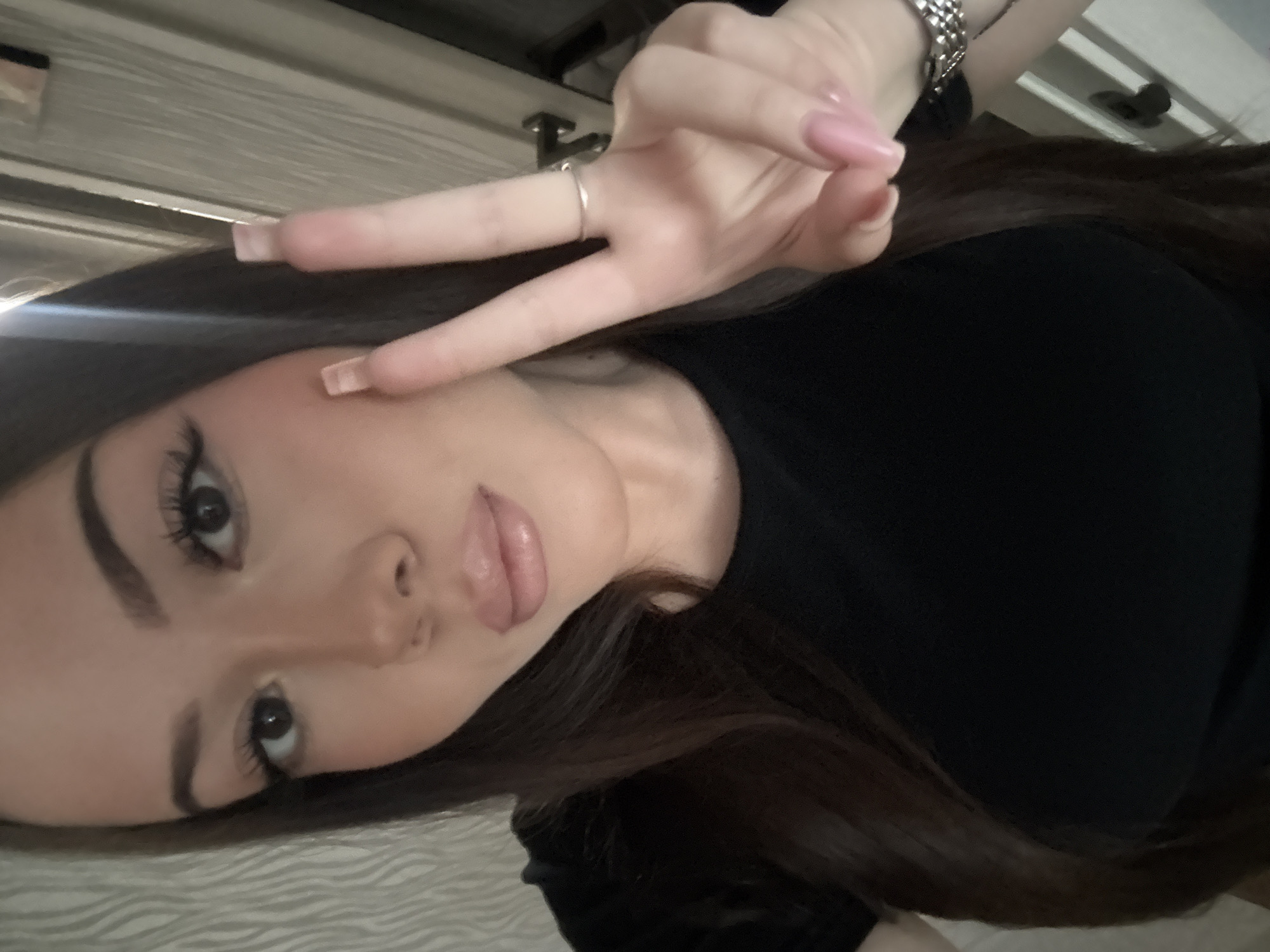 The image is a close-up, tilted selfie of a young woman, captured indoors. She is positioned diagonally across the frame, looking towards the camera with a calm expression.
The subject has long, dark brown hair, styled to fall around her face. She is wearing a plain black, short-sleeved top. Her face is made up, featuring dark, well-defined eyebrows, prominent eyelashes enhanced with mascara and eyeliner, and full lips adorned with a light pink or nude-colored lip gloss or lipstick.
Her right hand is raised into the upper left quadrant of the frame, forming a "peace" sign (V-sign) with her index and middle fingers. Her nails are long, manicured, and painted a light pink or nude shade. A simple silver-toned ring is visible on her index finger, and a silver metal-link watch is worn on her wrist.
The background is softly blurred, showing light-colored indoor surfaces with horizontal patterns, possibly a ceiling or wall, suggesting an interior setting. A bright light source or reflection is noticeable on the left side of the image. There is no visible text. The photograph appears to be an informal personal shot, taken in Naberezhnye Chelny, Russia.