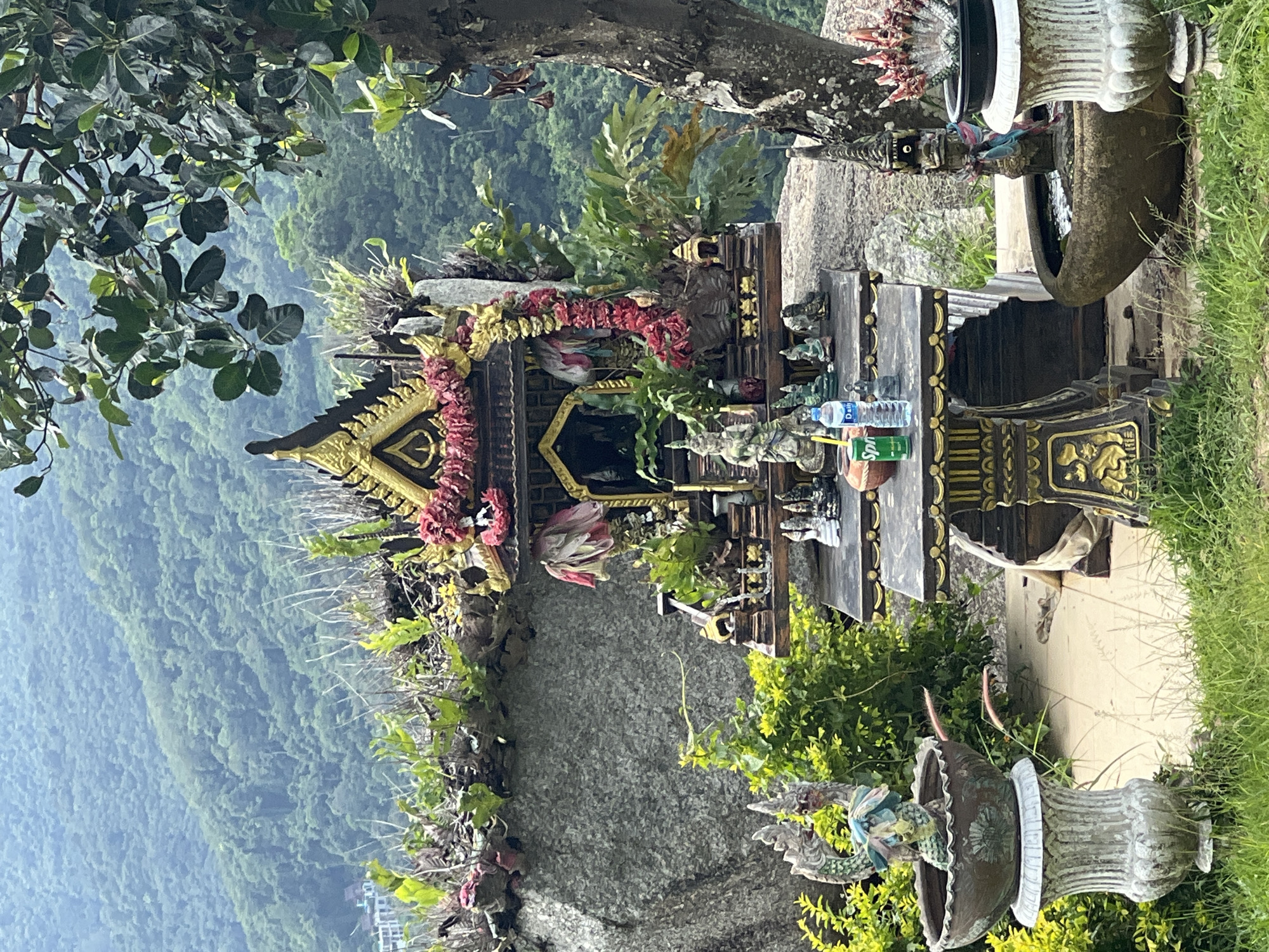 The image shows an outdoor spirit house, likely a San Phra Phum, located on a rocky hillside with lush green trees and vegetation in the background, characteristic of Karon, Thailand. The spirit house itself is ornate, with a multi-tiered, dark wood structure adorned with gold trim and decorative red and pink floral garlands. Several small statues, possibly deities or guardians, are placed on and around the spirit house. In front of the spirit house is a table with more offerings, including two bottles of drinks (a Sprite can and a bottle of water) and various small figurines. Flanking the spirit house are two large, decorative urns. The urn on the left holds a statue of a dragon-like creature, while the urn on the right appears to contain water and a decorative element. The overall scene suggests a place of worship or veneration, situated in a natural, elevated environment with a distant view of a densely forested mountainside, possibly overlooking a coastal area as implied by the location in Karon. There are no people visible in the image.